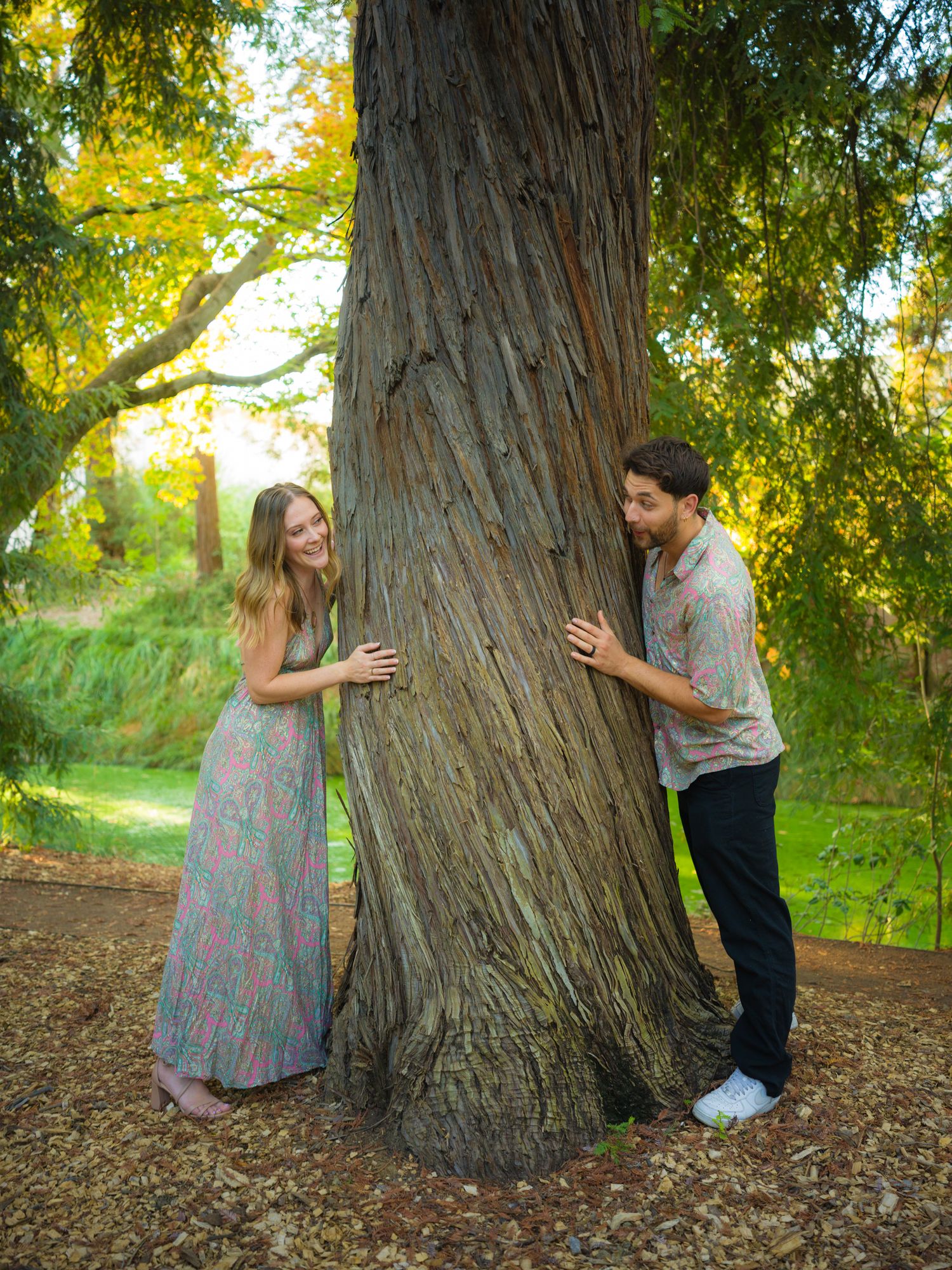Couple plays peek-a-boo around large tree trunk during outdoor photo shoot.
