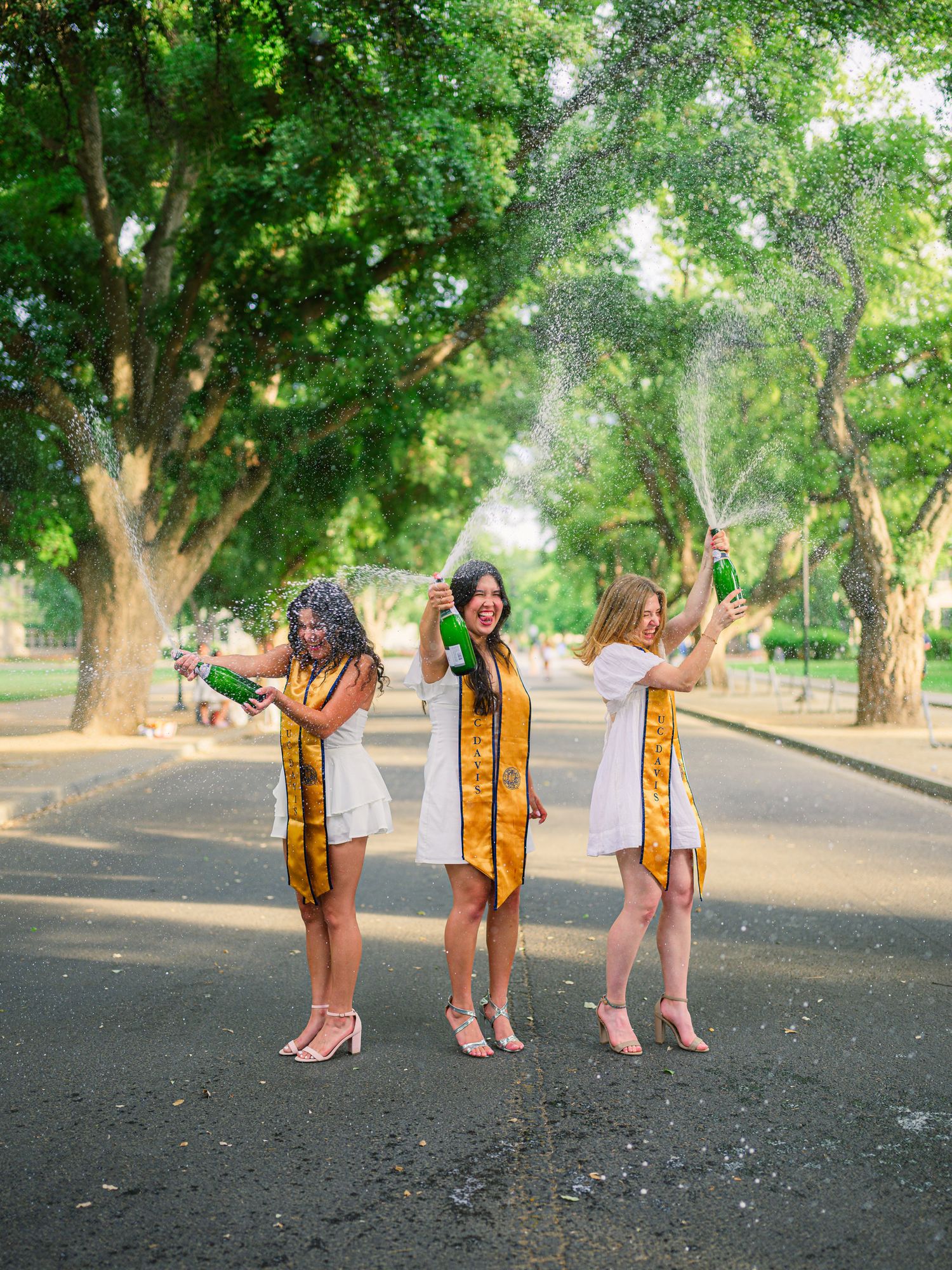 Three graduates in yellow stoles pose playfully in a tree-lined outdoor setting.