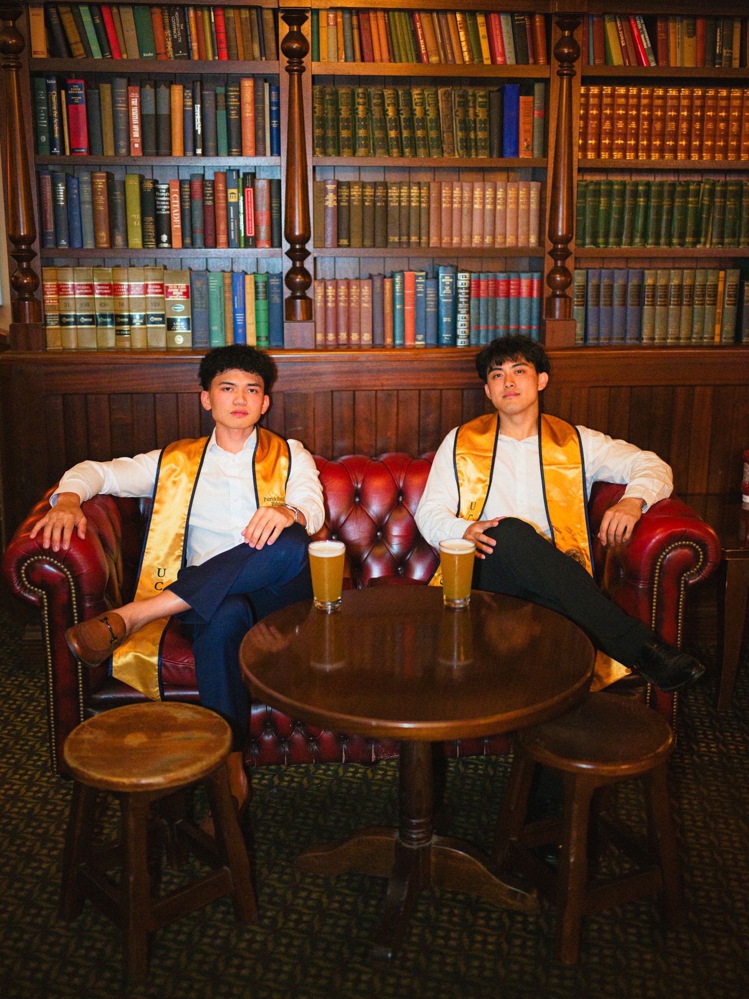 Two graduates in gold stoles sit on red leather couch in library with wooden bookshelves.