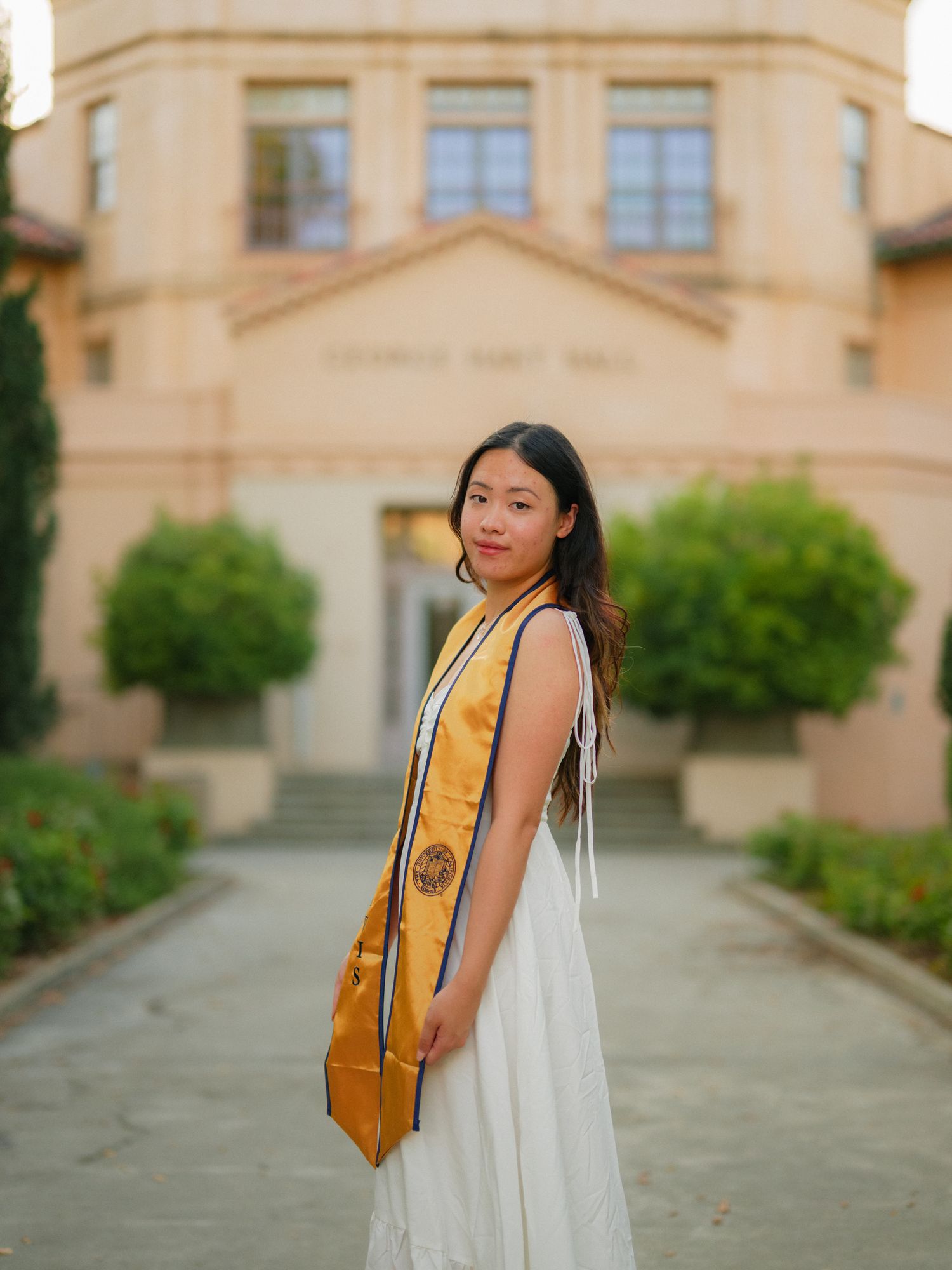 A graduate in a white dress and gold sash stands in front of a stately building entrance with manicured landscaping.