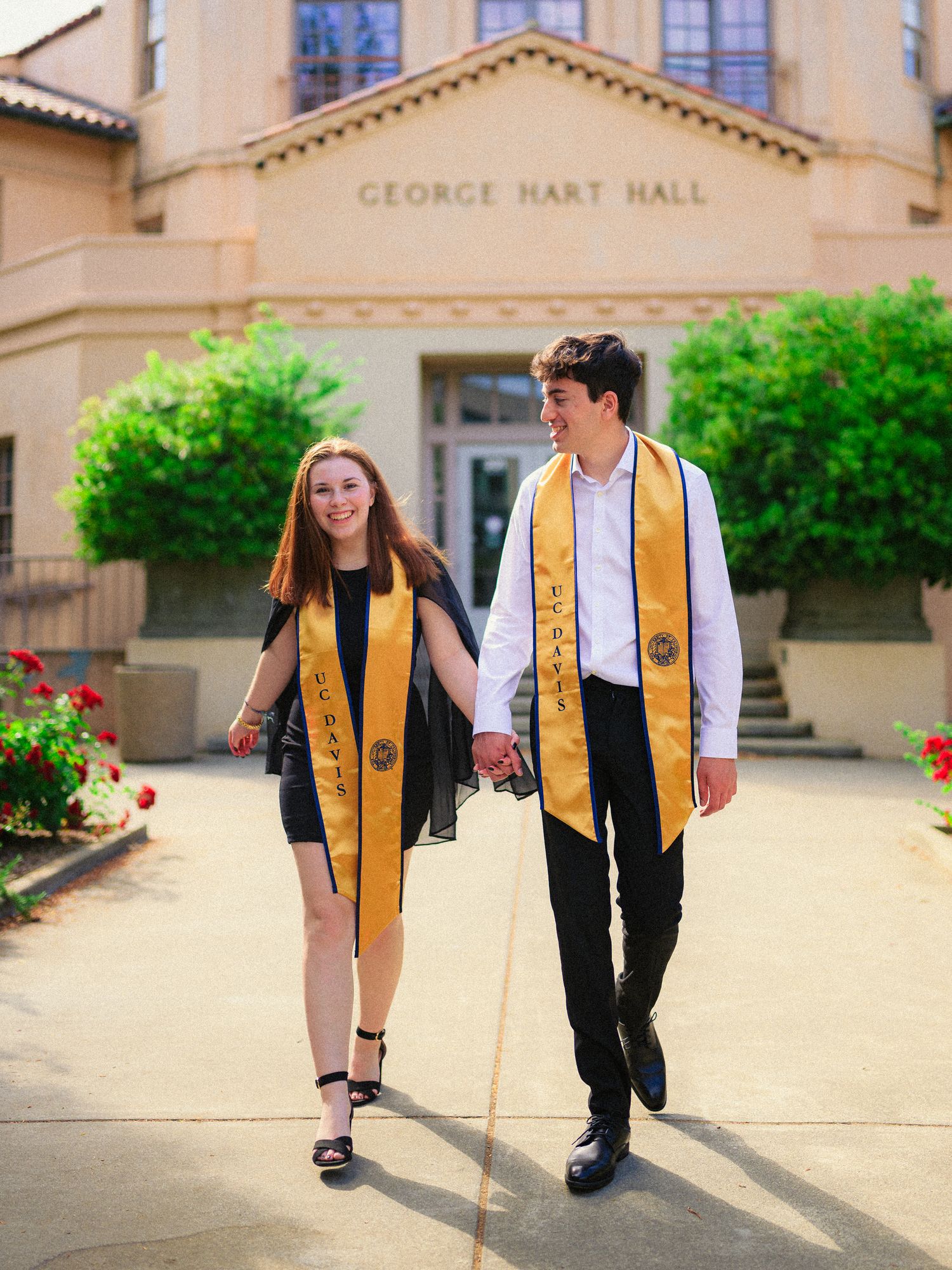 Graduates walk hand in hand outside George Hart Hall wearing matching gold graduation stoles.