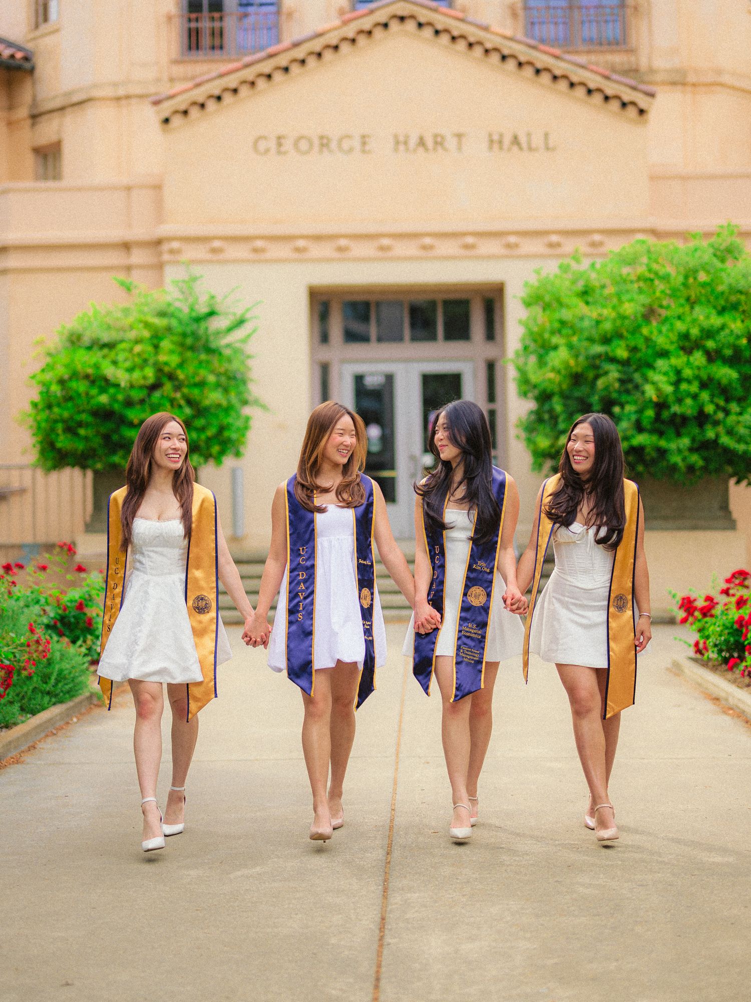 A group of friends in white dresses walking together along a stone building pathway with green bushes and flowers.