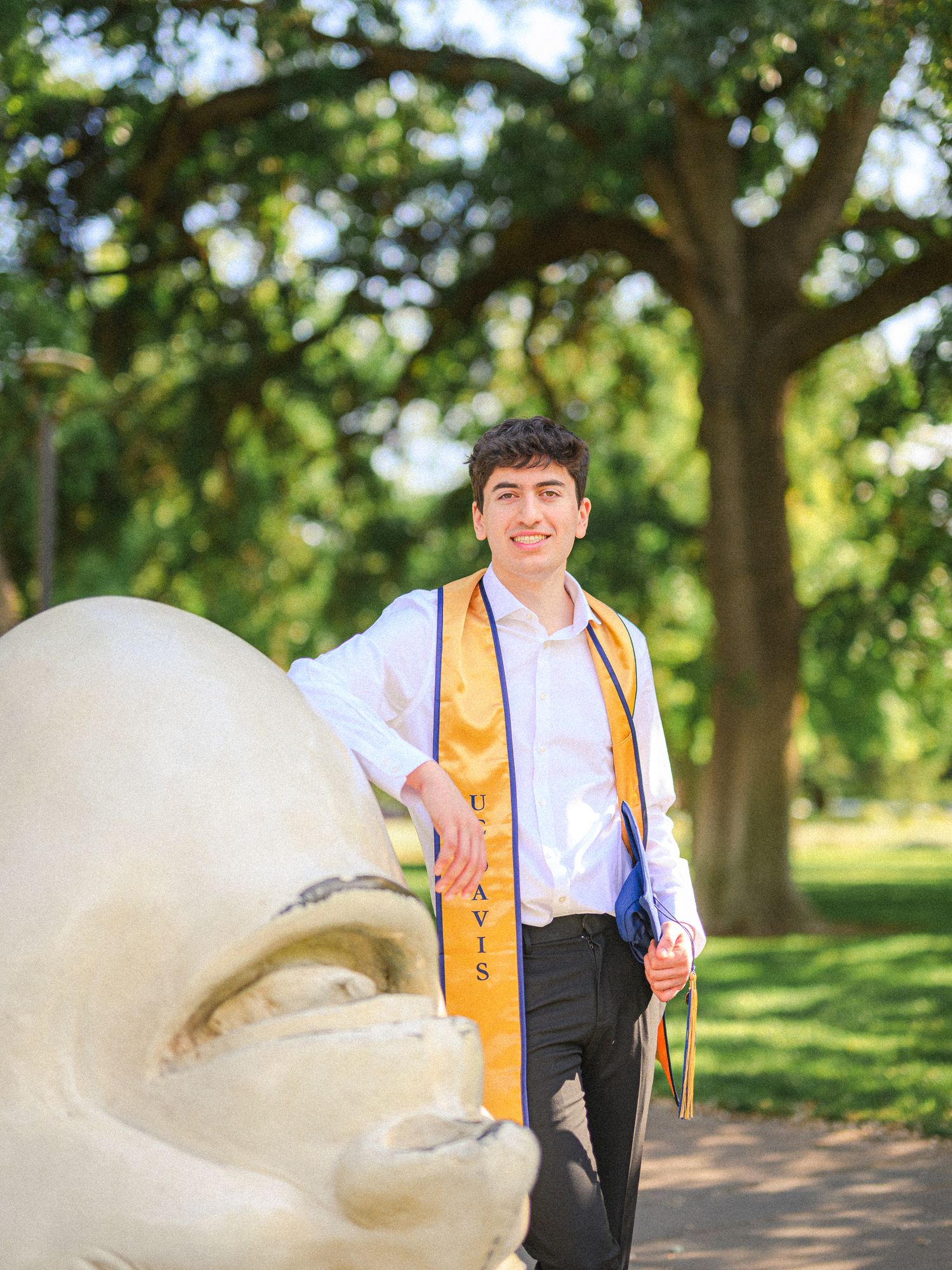 Graduate in cap and gown with yellow stole walking along a sunny pathway surrounded by trees.