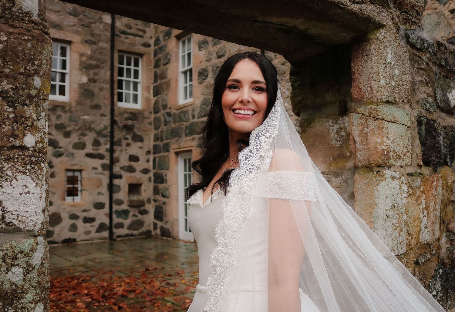 A bride in a white wedding dress with lace details stands in front of a rustic stone wall on her wedding day.