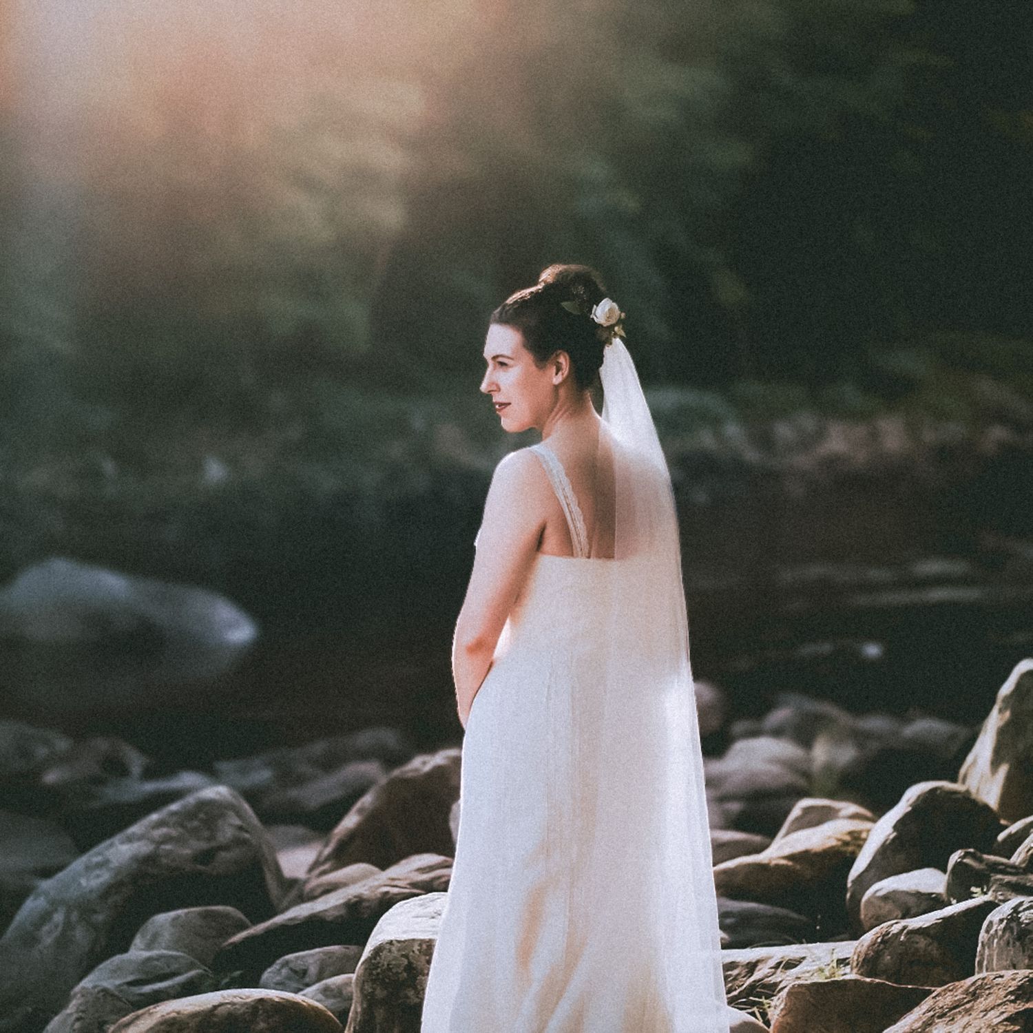 Bride in flowing white dress and long veil stands on rocky riverbank at sunset in moody outdoor setting.