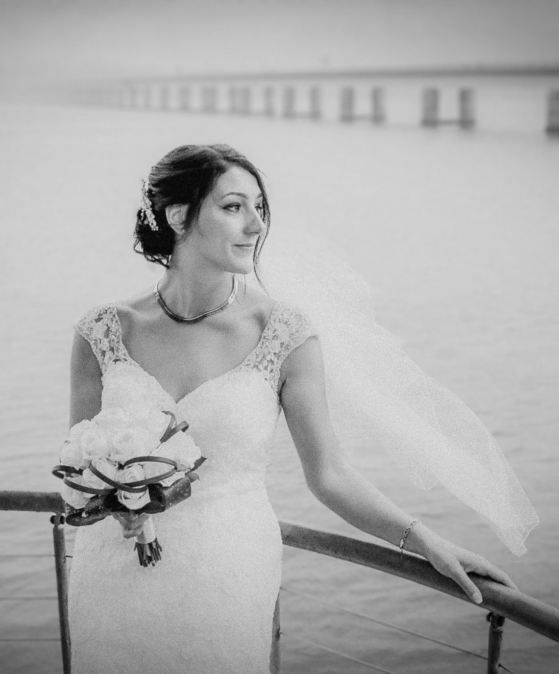 A bride in a lace wedding dress holds a bouquet while standing near a waterfront bridge in black and white.