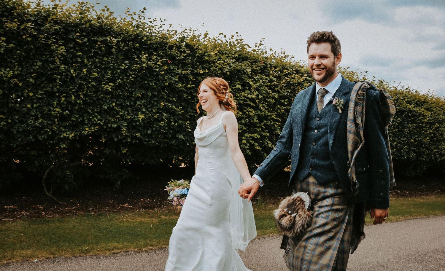 A couple walks together on a country path surrounded by lush green hedgerows on their wedding day.
