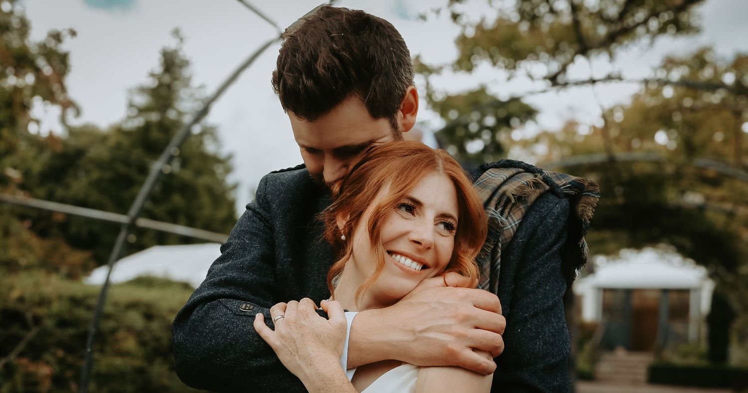A couple embraces outdoors in a romantic moment during golden hour with natural lighting and greenery in background.
