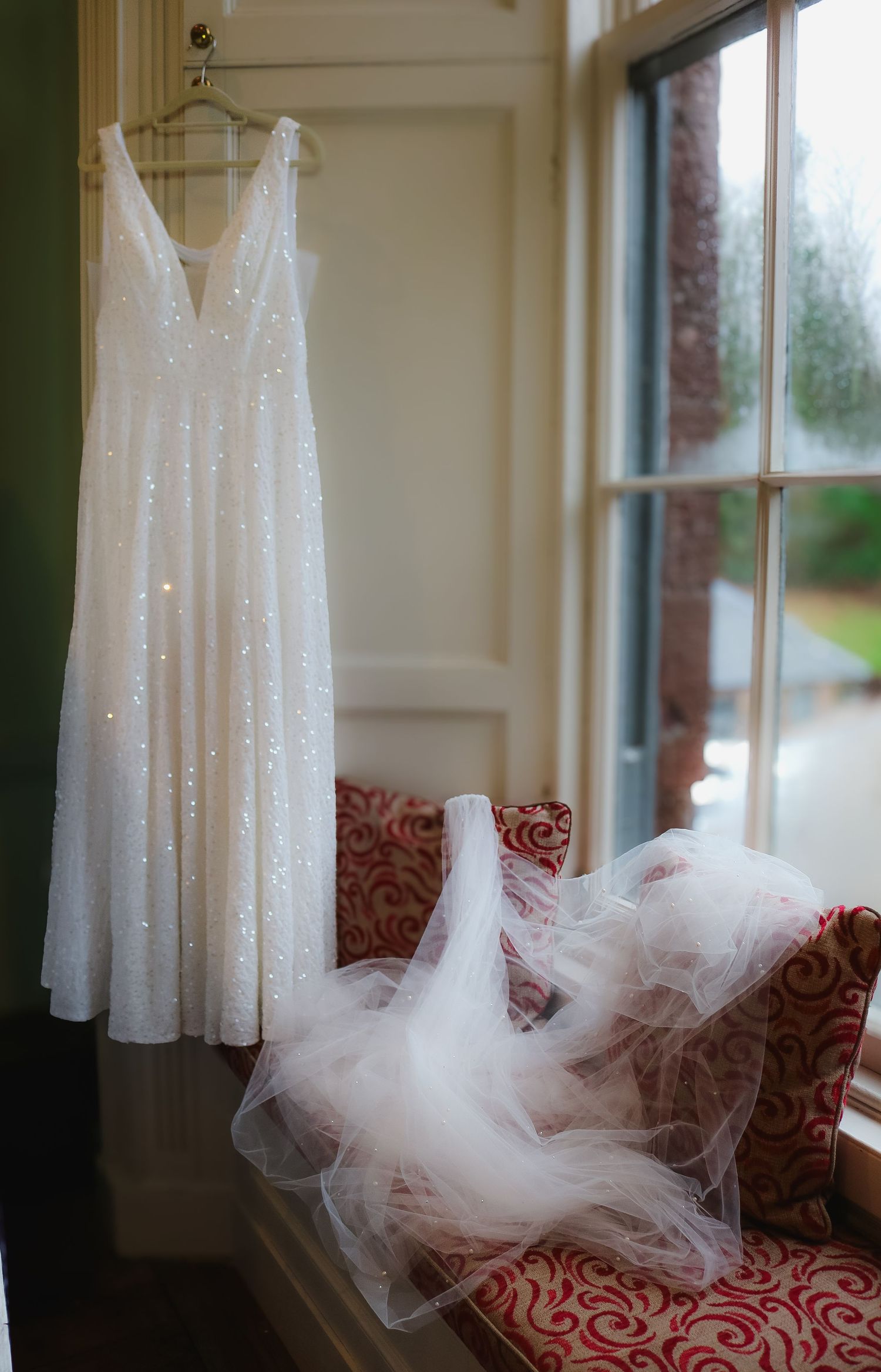 A white sequined wedding dress hangs by a window with a delicate tulle veil draped on a red patterned chair.