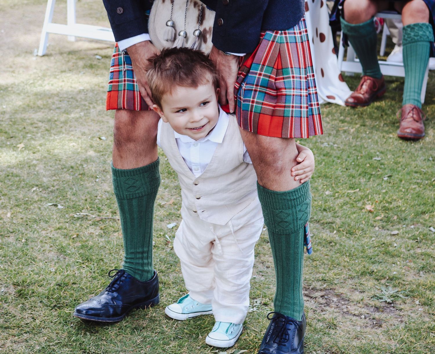 Small child in white outfit stands between adults wearing traditional Scottish kilts and green knee socks on grass.