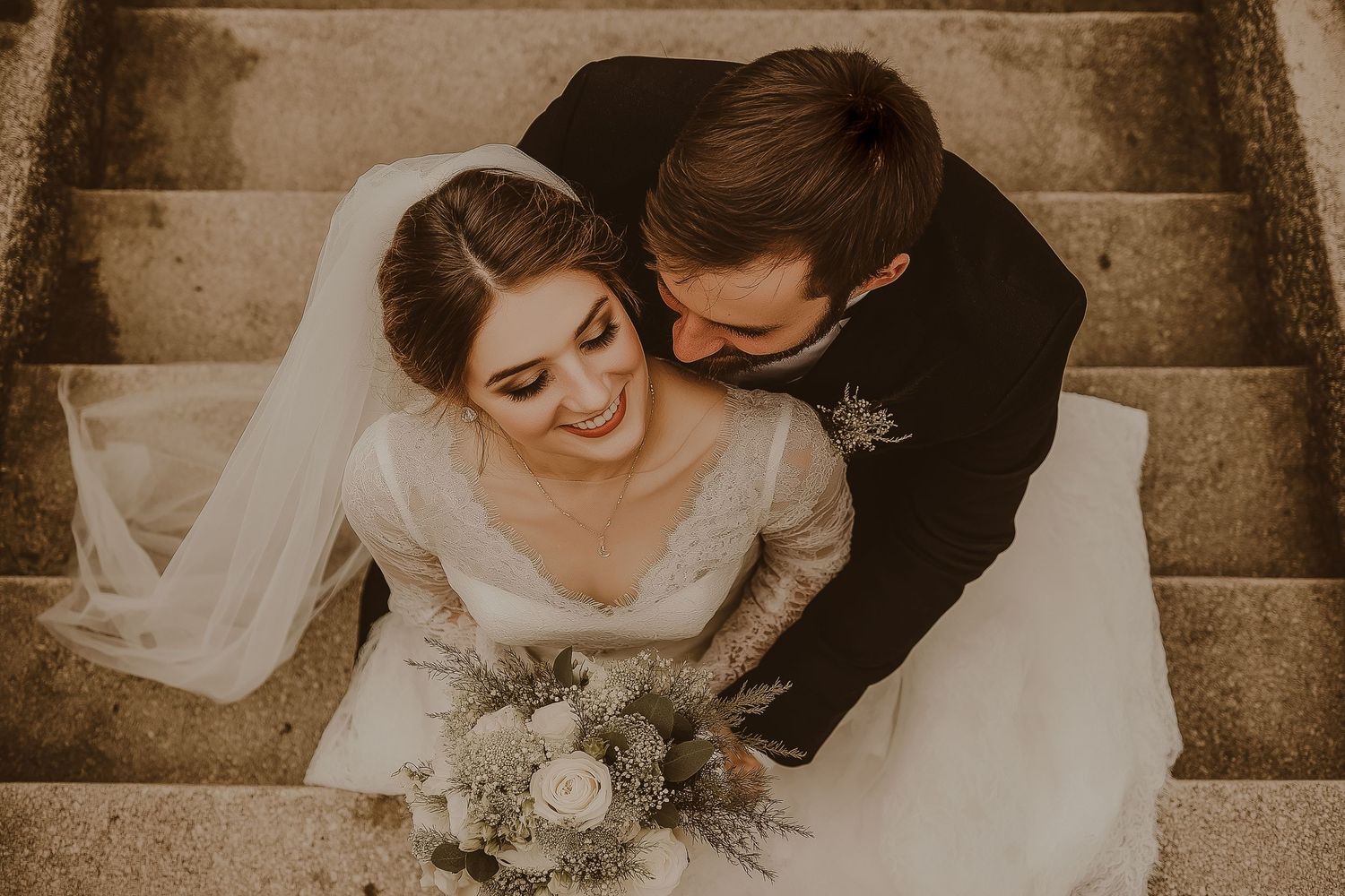 A romantic wedding couple sits on stone steps with a white bouquet in a warm sepia-toned photograph.