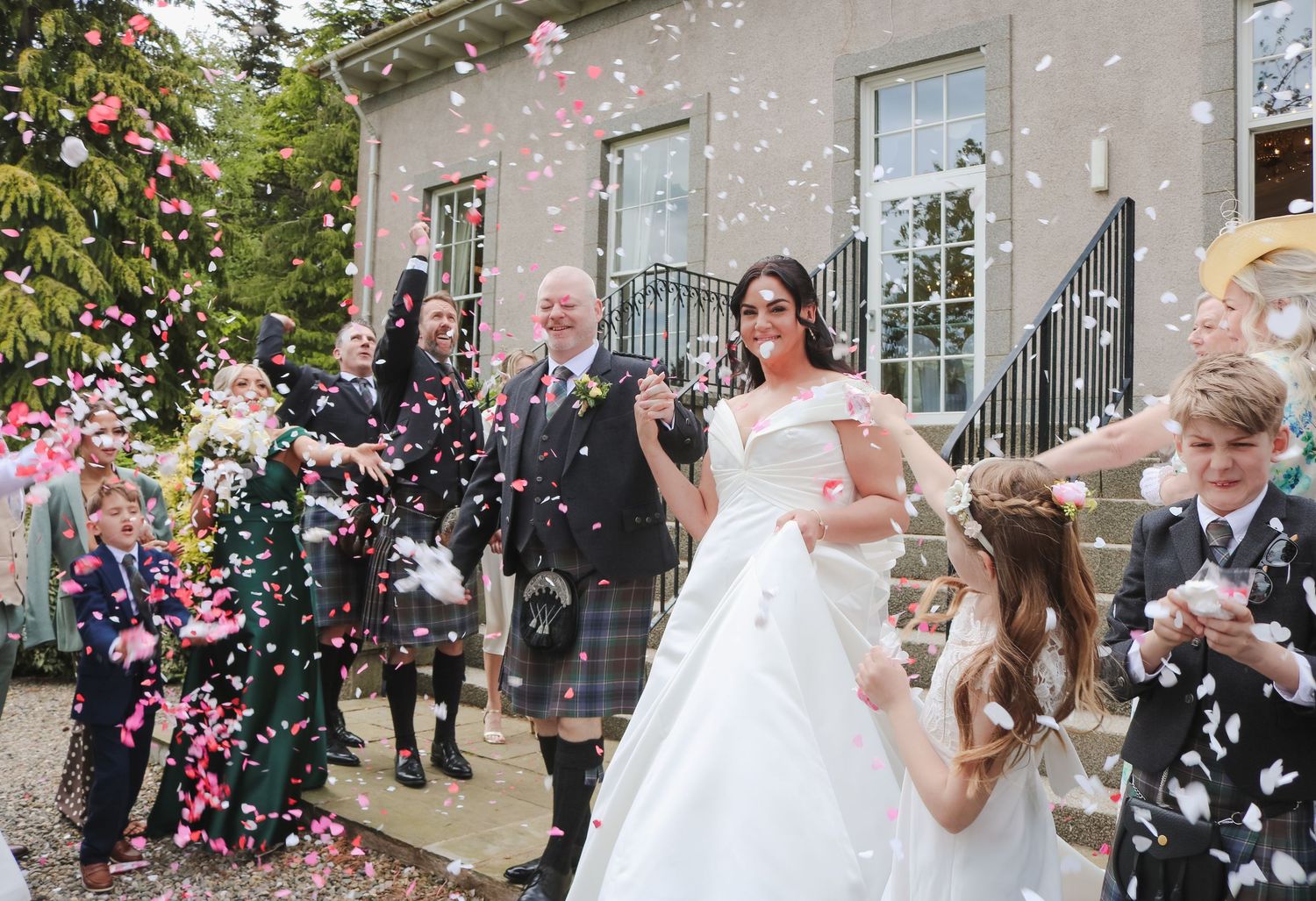 Bride and groom walk through colorful confetti shower outside stone venue surrounded by wedding guests in kilts.