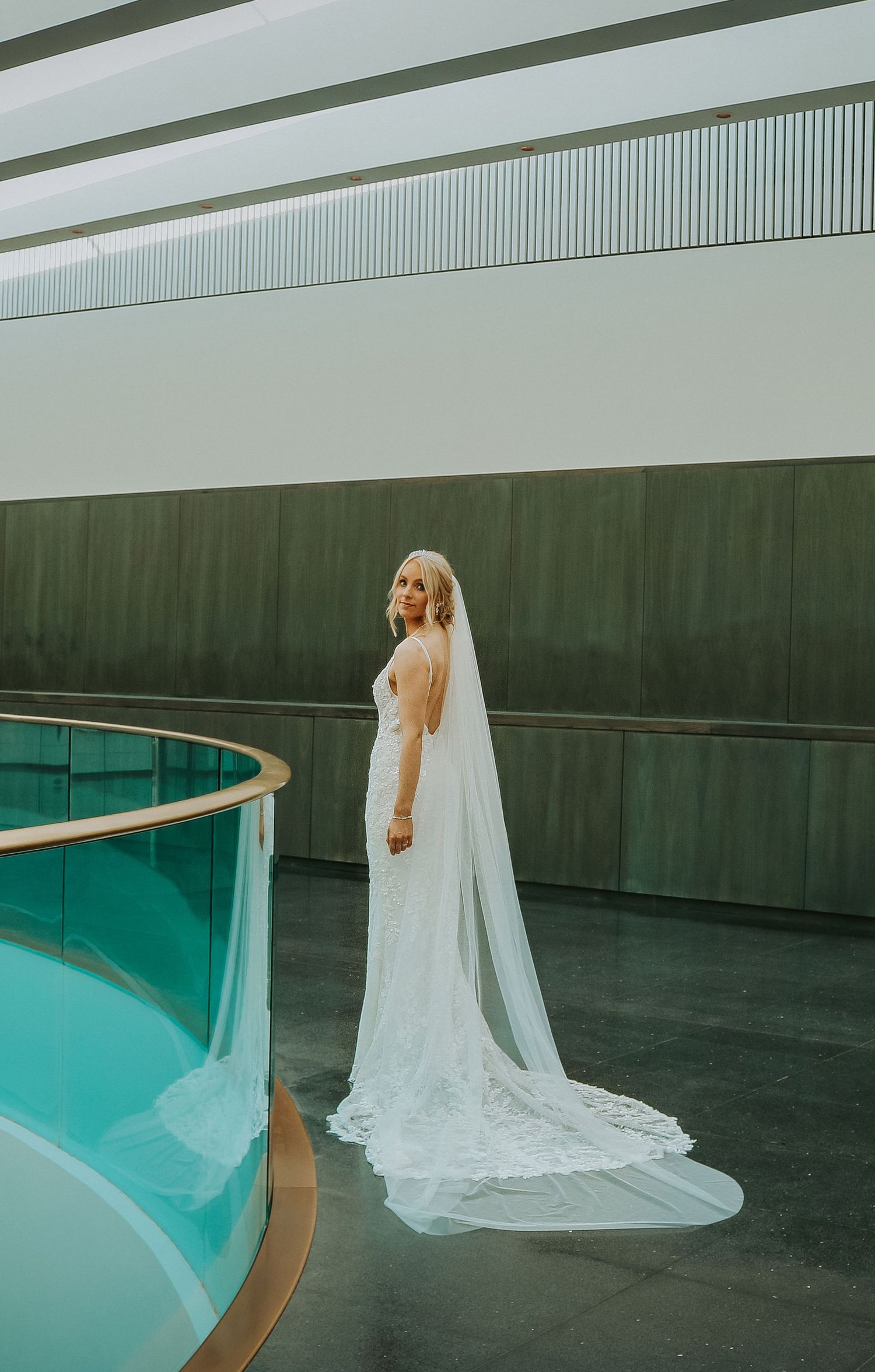 Elegant bride in a flowing white gown and long veil stands next to a curved turquoise pool in a modern architectural setting.
