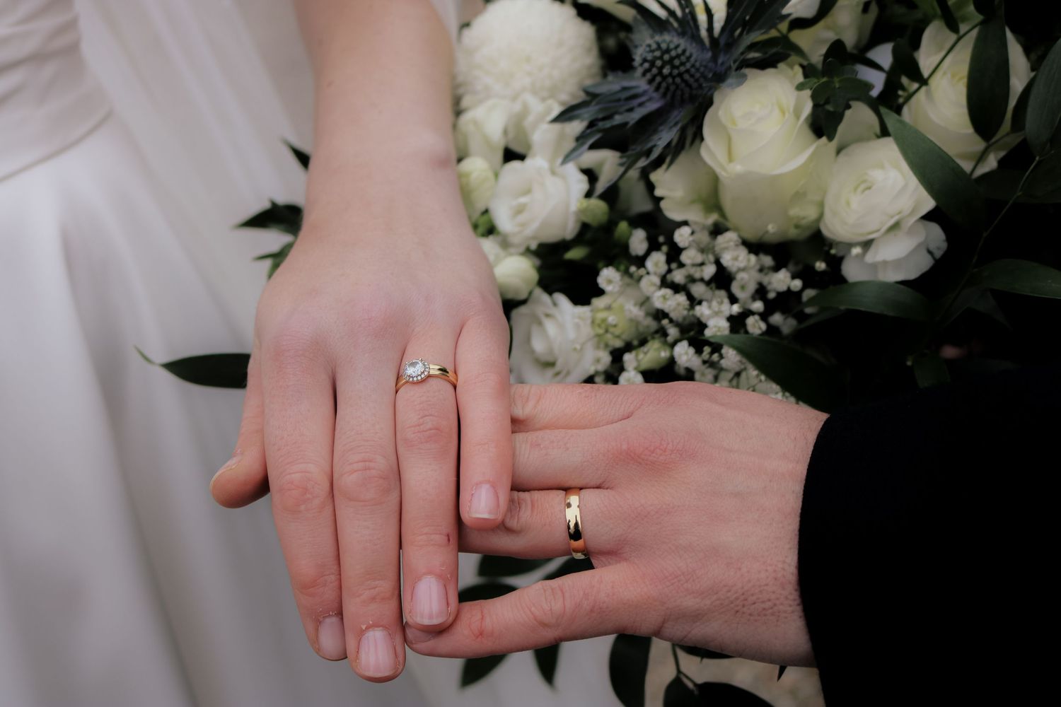 Two hands wearing wedding bands touch gently beside white peony and baby's breath bouquet.