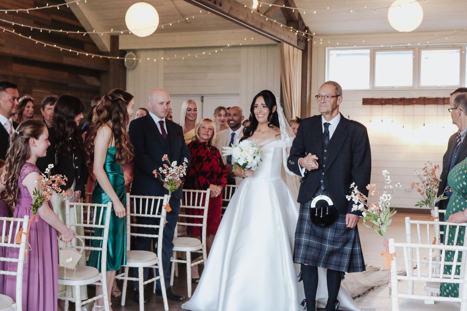 Wedding ceremony guests stand in a row during a procession at an indoor venue decorated with string lights and white chairs.