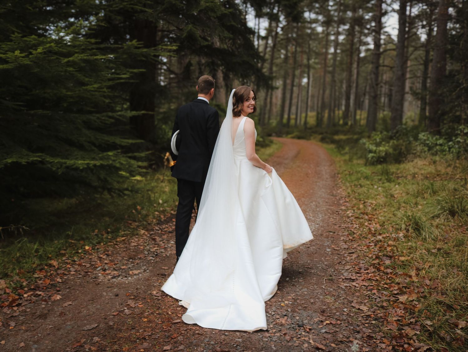 A couple in wedding attire walk along a wooded forest path covered in autumn leaves.