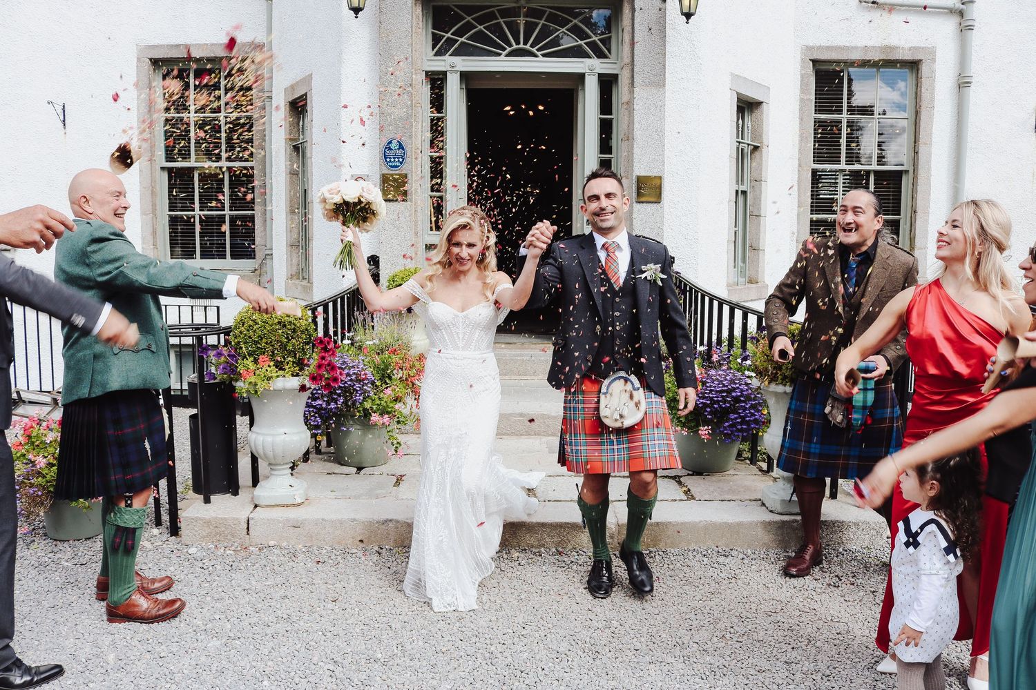 Wedding guests throw rice and petals in celebration outside a historic stone building entrance.
