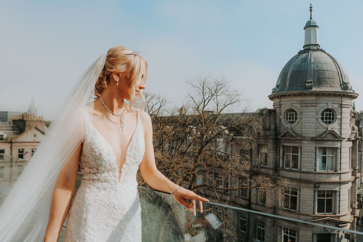 A bride in a lace gown stands on a balcony overlooking historic architecture with a domed building.