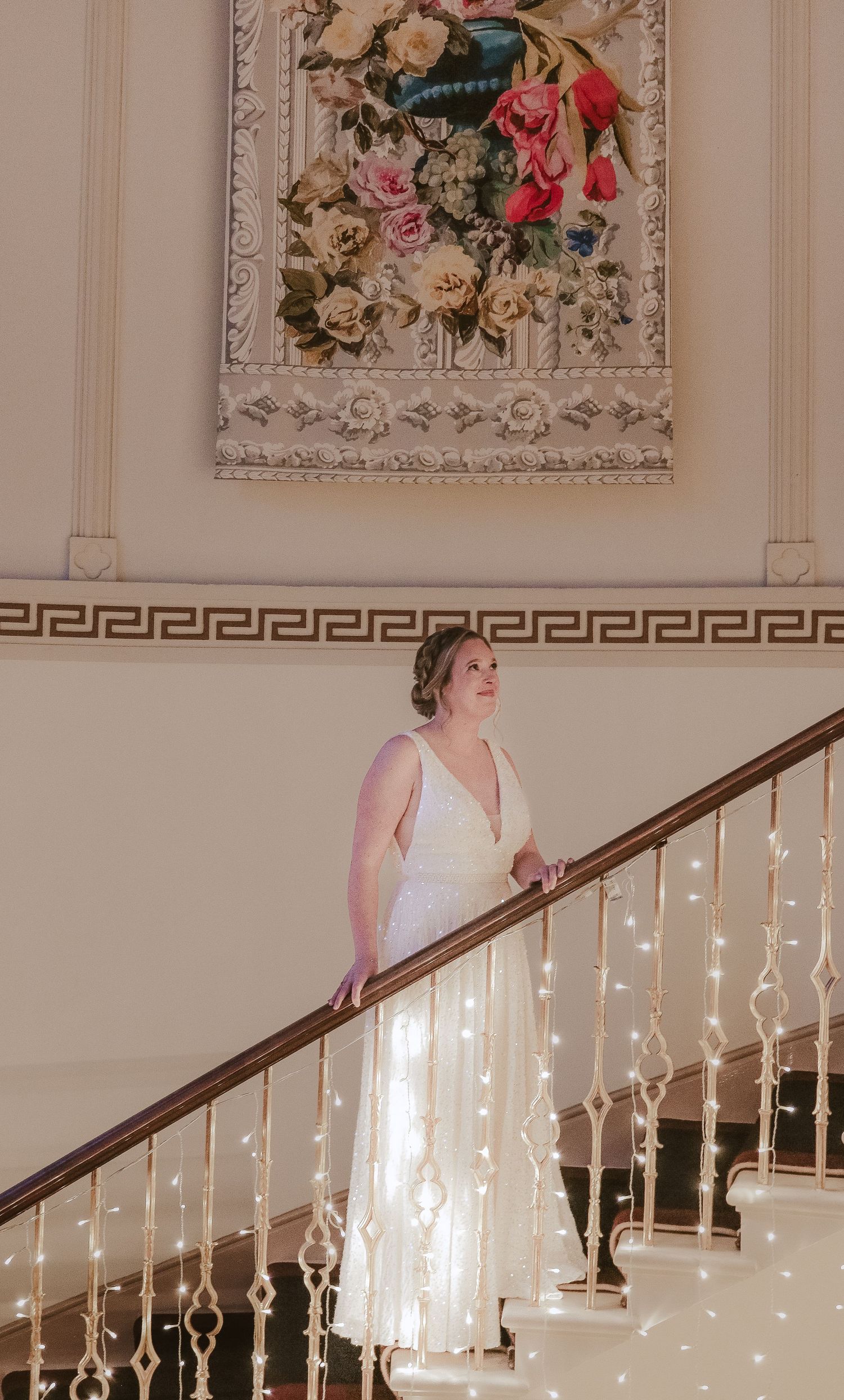 Elegant bride in white lace dress descends ornate staircase decorated with glowing fairy lights beneath vintage wall art decor.