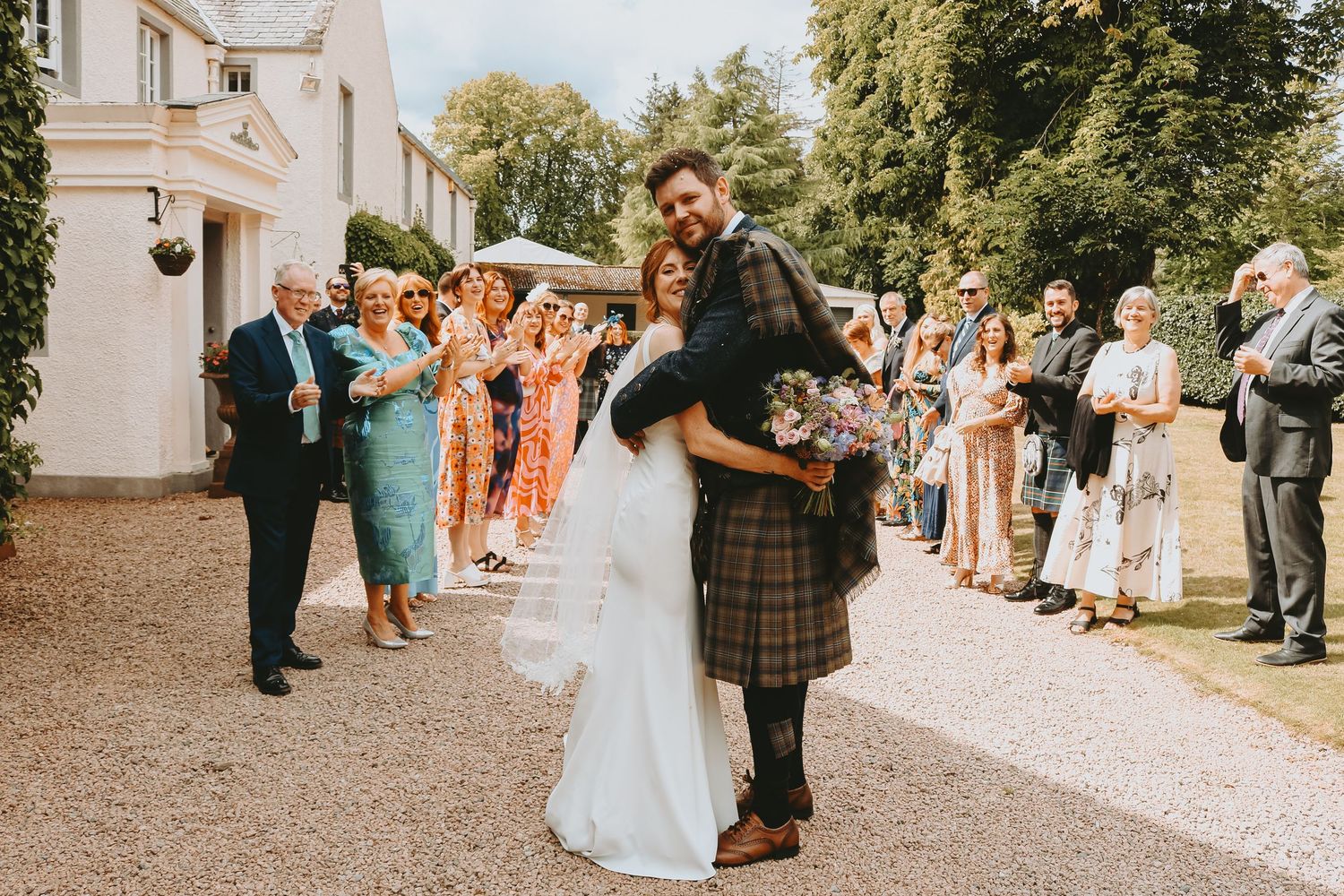 Wedding guests line up outside a venue to celebrate the newlyweds with bubbles and confetti on a sunny day.