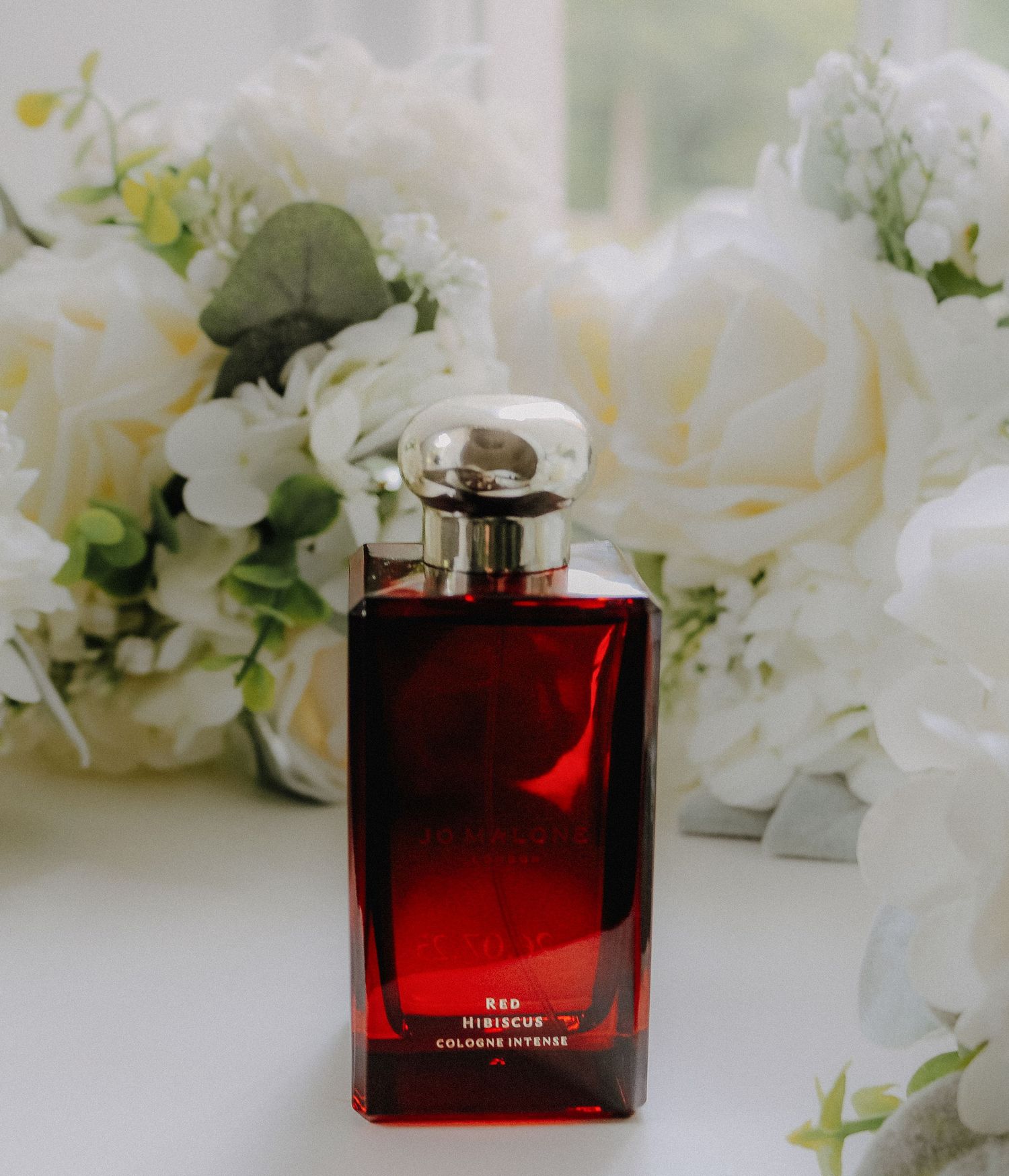 Red perfume bottle displayed on white floral background with roses and delicate petals.