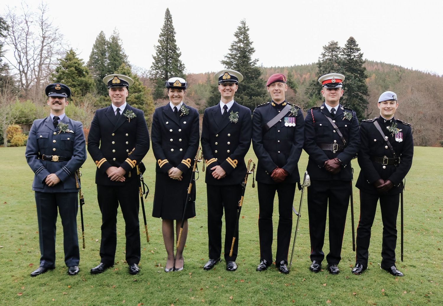 A line of police officers in dress uniforms stand at attention outdoors with evergreen trees behind them.