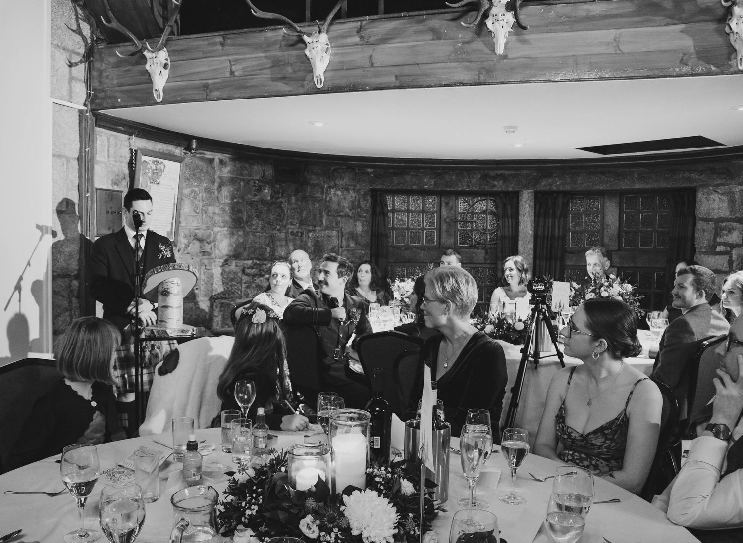 A black and white photo of guests gathered at an elegant dinner reception with floral centerpieces and formal attire.