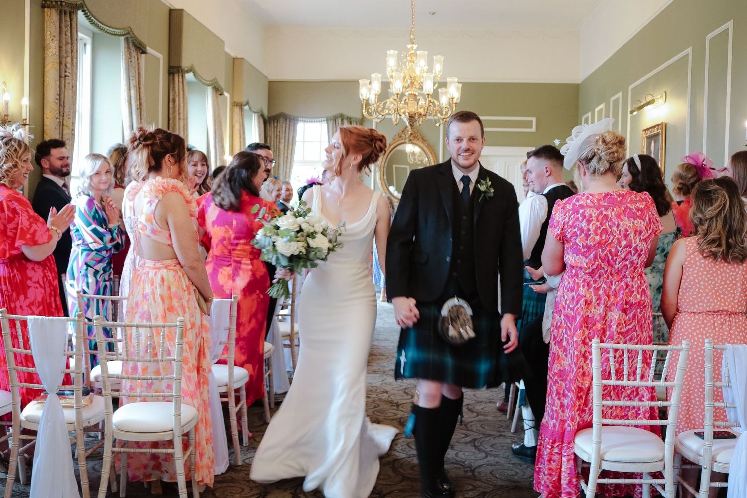 Elegant indoor wedding ceremony with guests in pink dresses and traditional Scottish attire beneath a crystal chandelier.