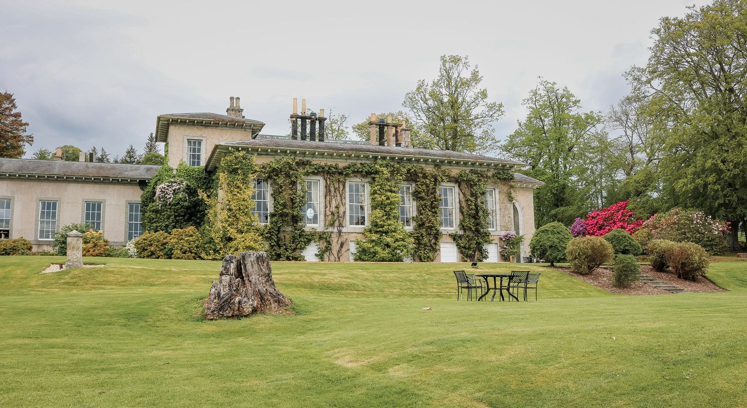 Ivy-covered country manor house with manicured lawn, outdoor seating, and colorful garden shrubs under cloudy skies.