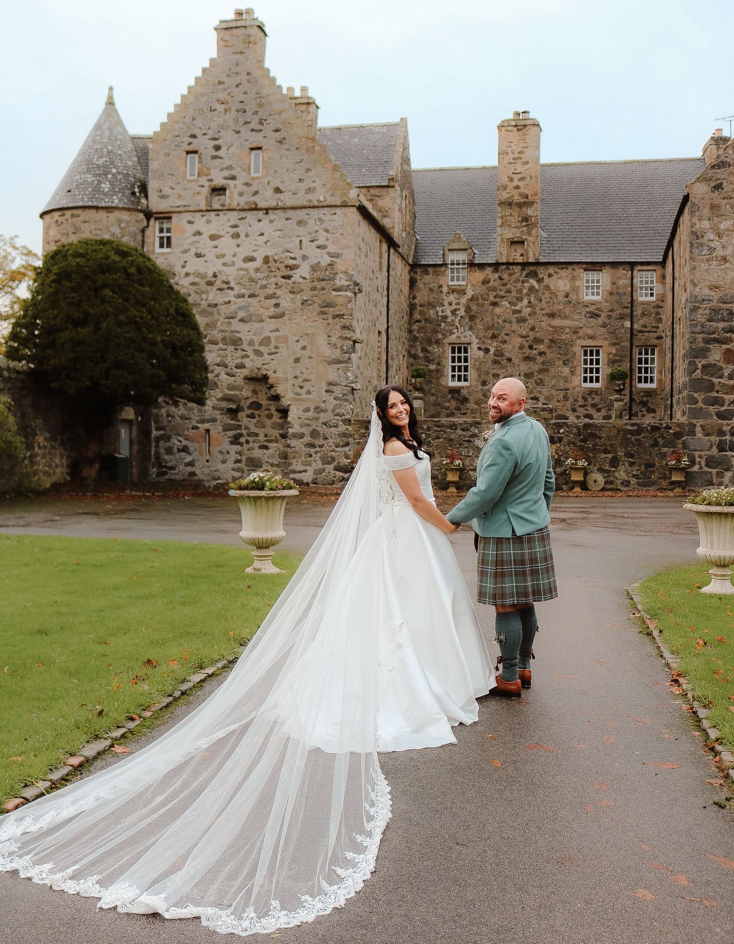 A couple in wedding attire walk along a stone path in front of a historic Scottish castle on their wedding day.