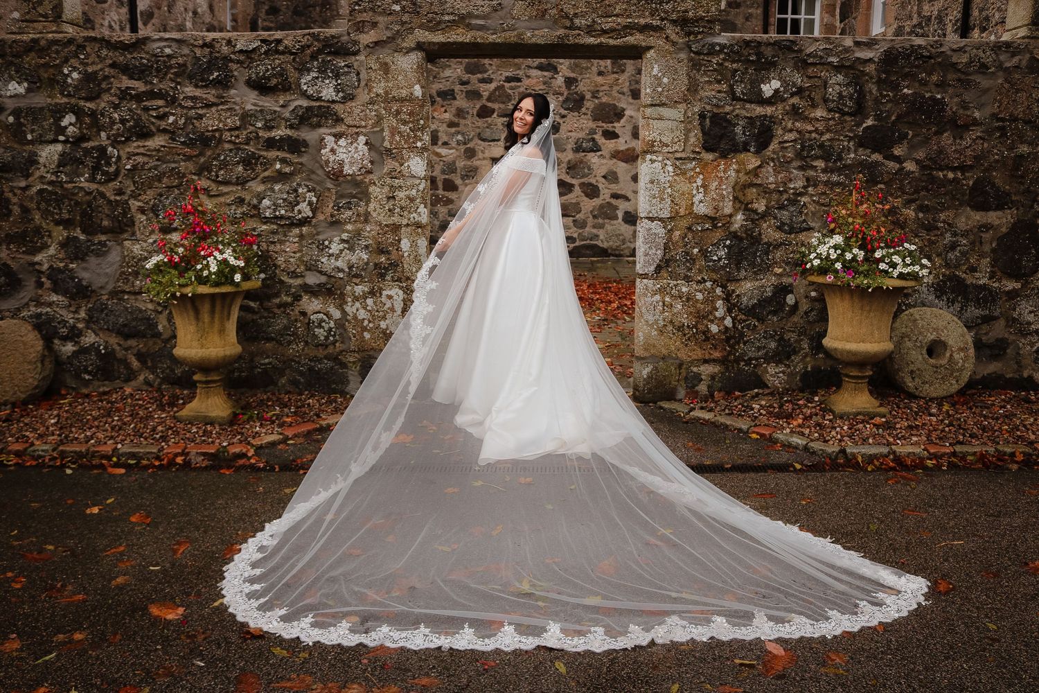 A long white wedding veil trails behind on stone steps surrounded by autumn leaves and stone walls.