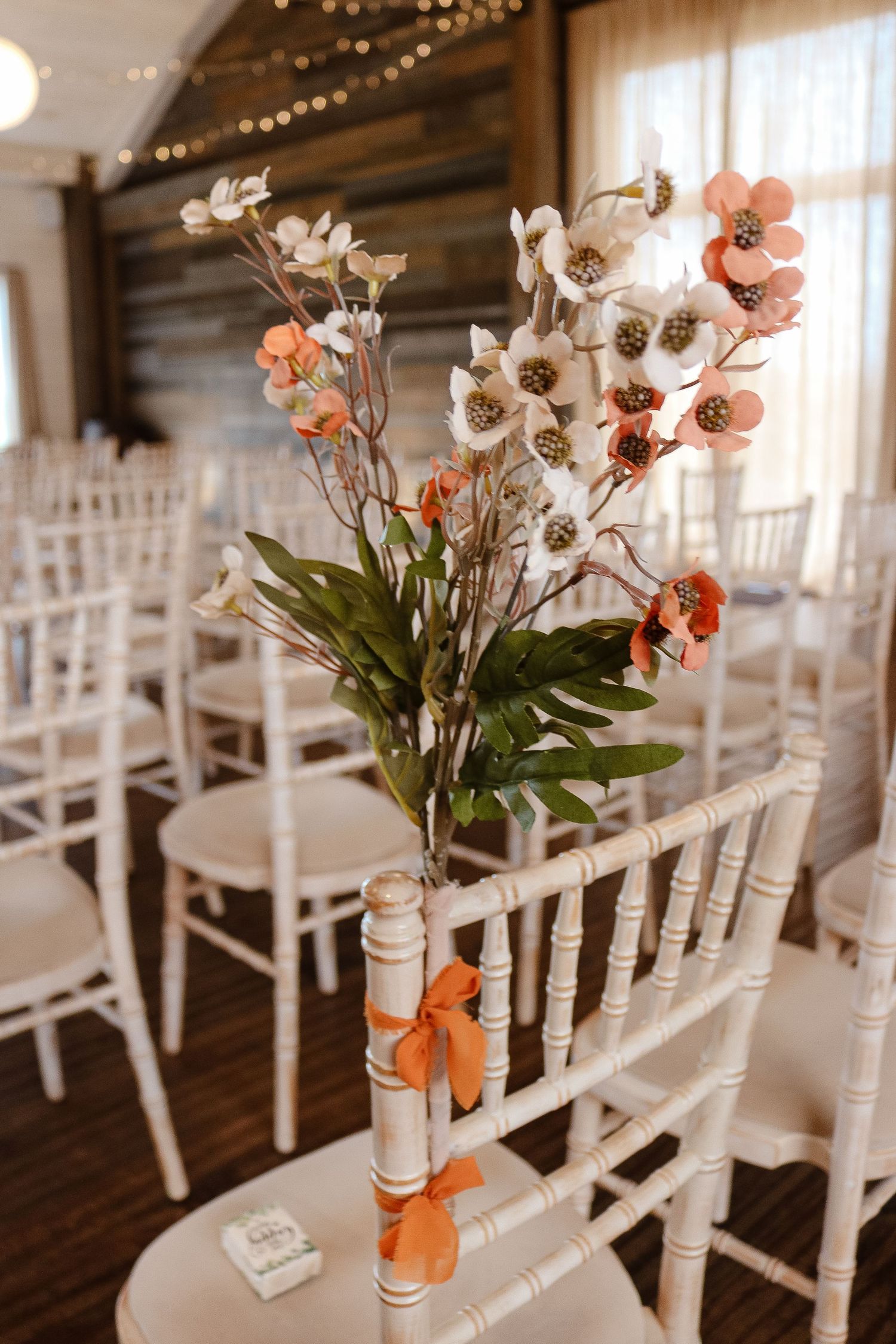 White chairs adorned with peach and cream floral arrangements line up for an elegant indoor wedding ceremony.