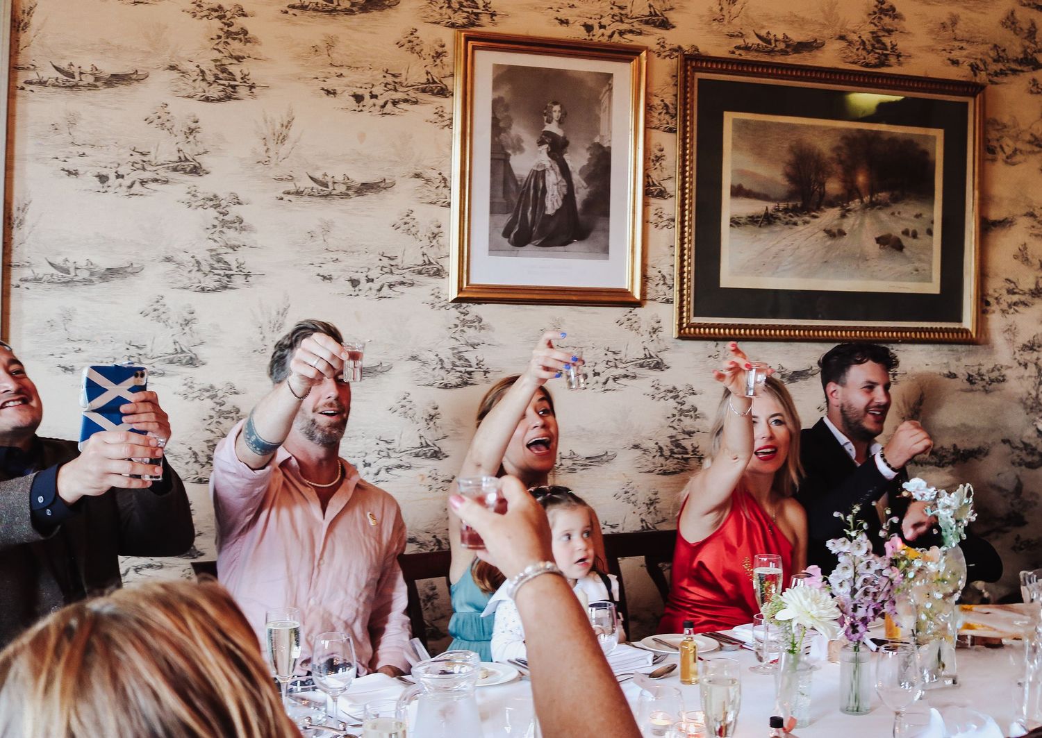 Wedding guests all take shots together at a reception with decorative wallpaper and framed artwork in the background.