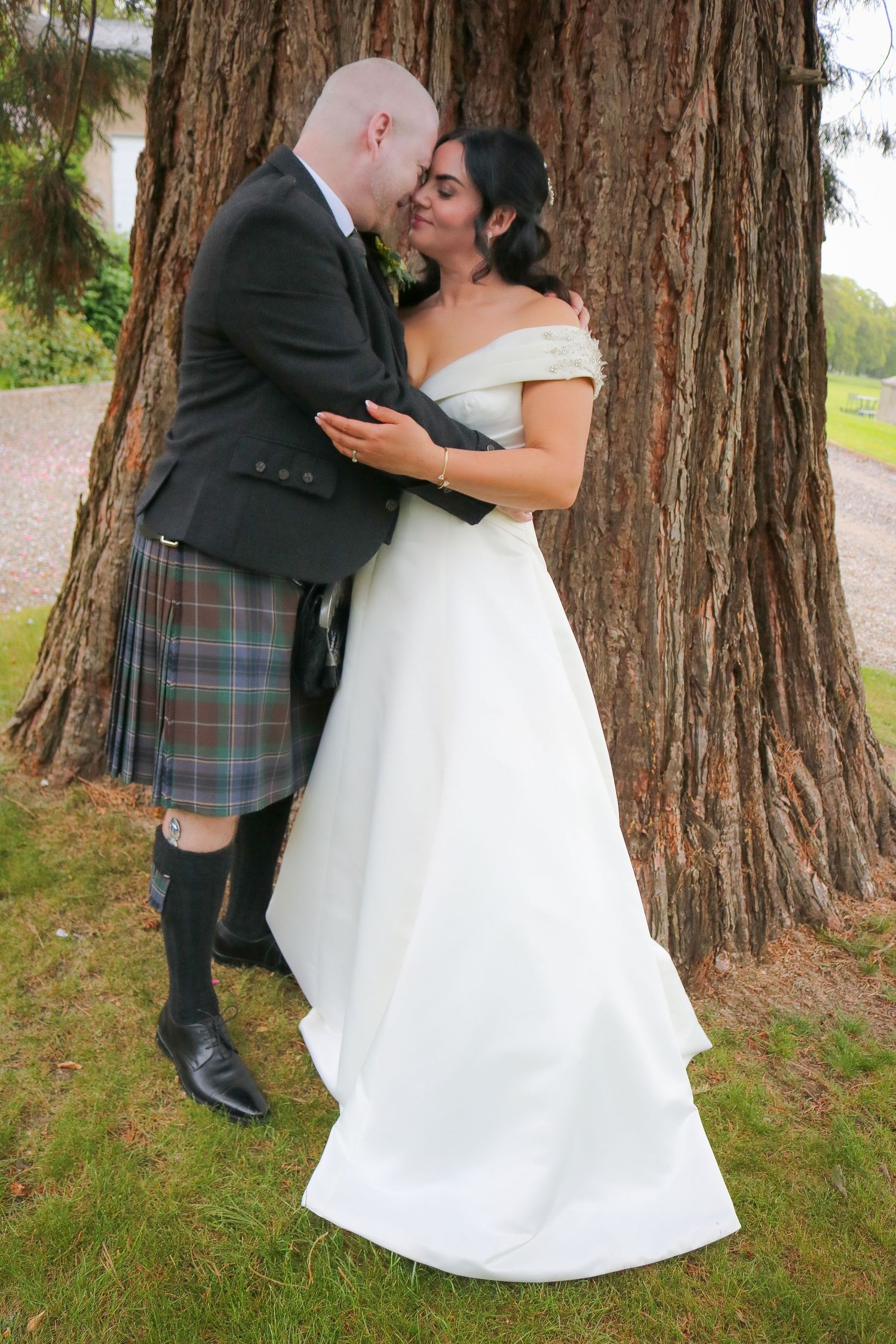 A bride in a white off-shoulder gown and groom in a tartan kilt share a romantic kiss against a large tree.