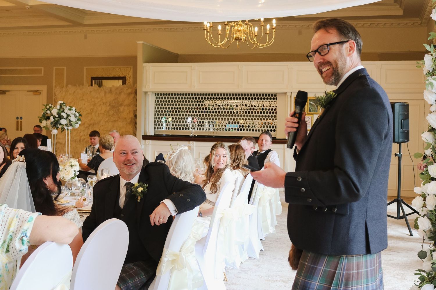 Man in kilt gives speech at elegant wedding reception, guests laughing and smiling in decorated venue.