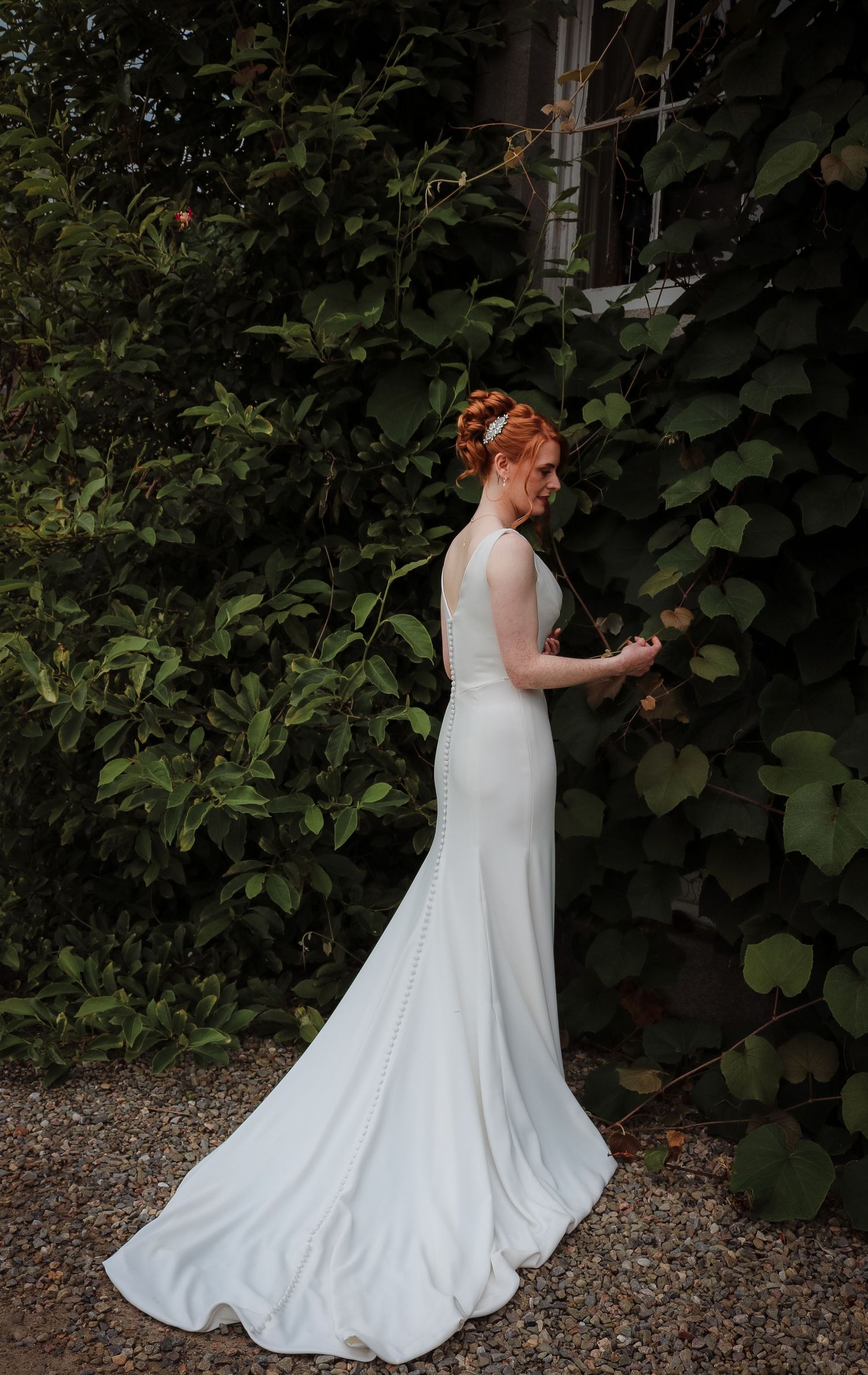 A bride in a flowing white wedding gown poses elegantly against a backdrop of lush green foliage in a garden setting.