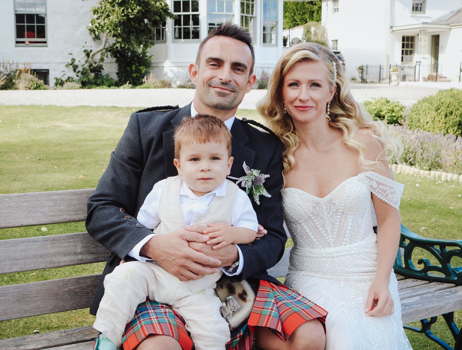A family sits together on a garden bench wearing formal attire including a kilt at an elegant white house.