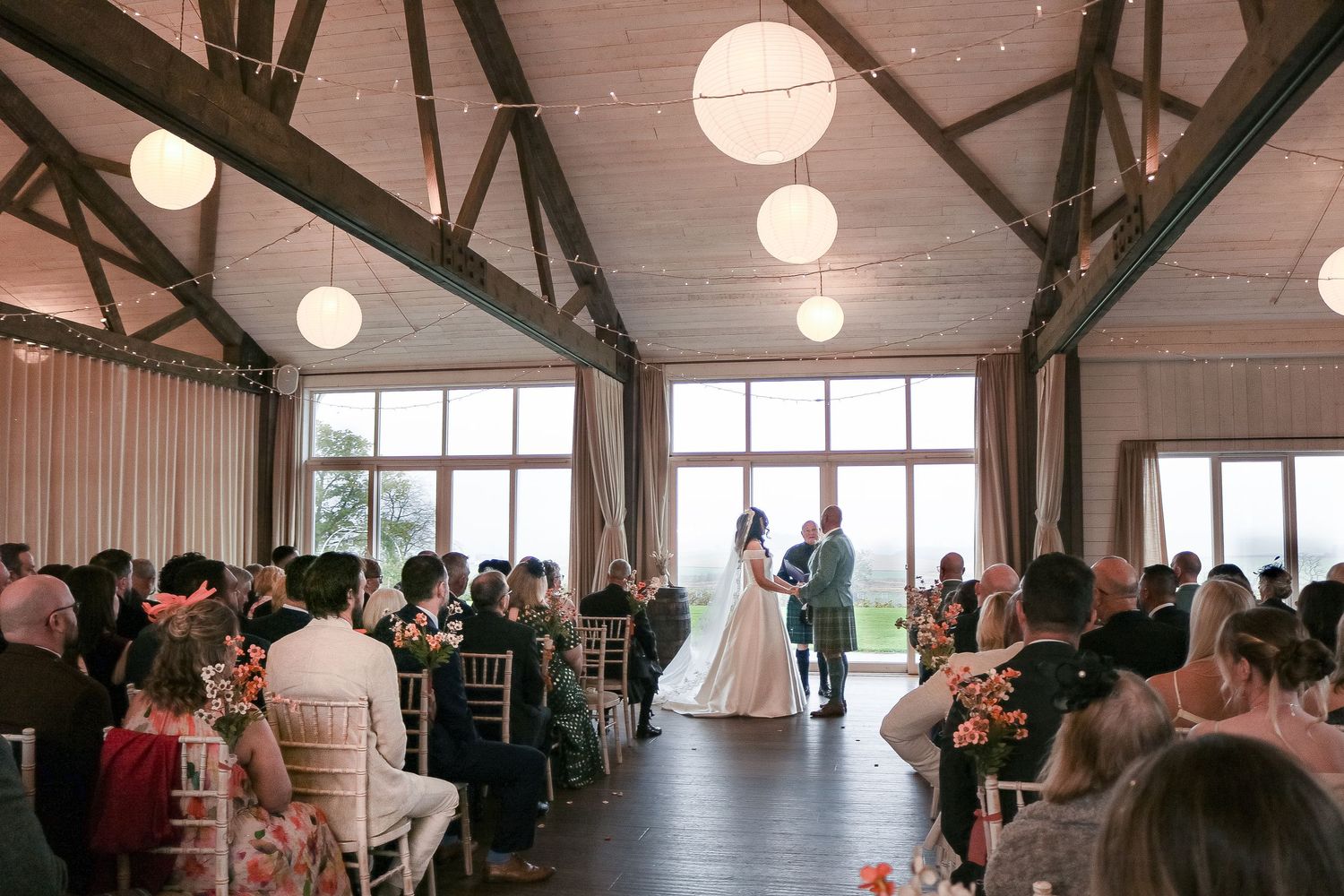 A wedding ceremony in a rustic venue with exposed beams, round paper lanterns, and guests seated in rows watching the couple.