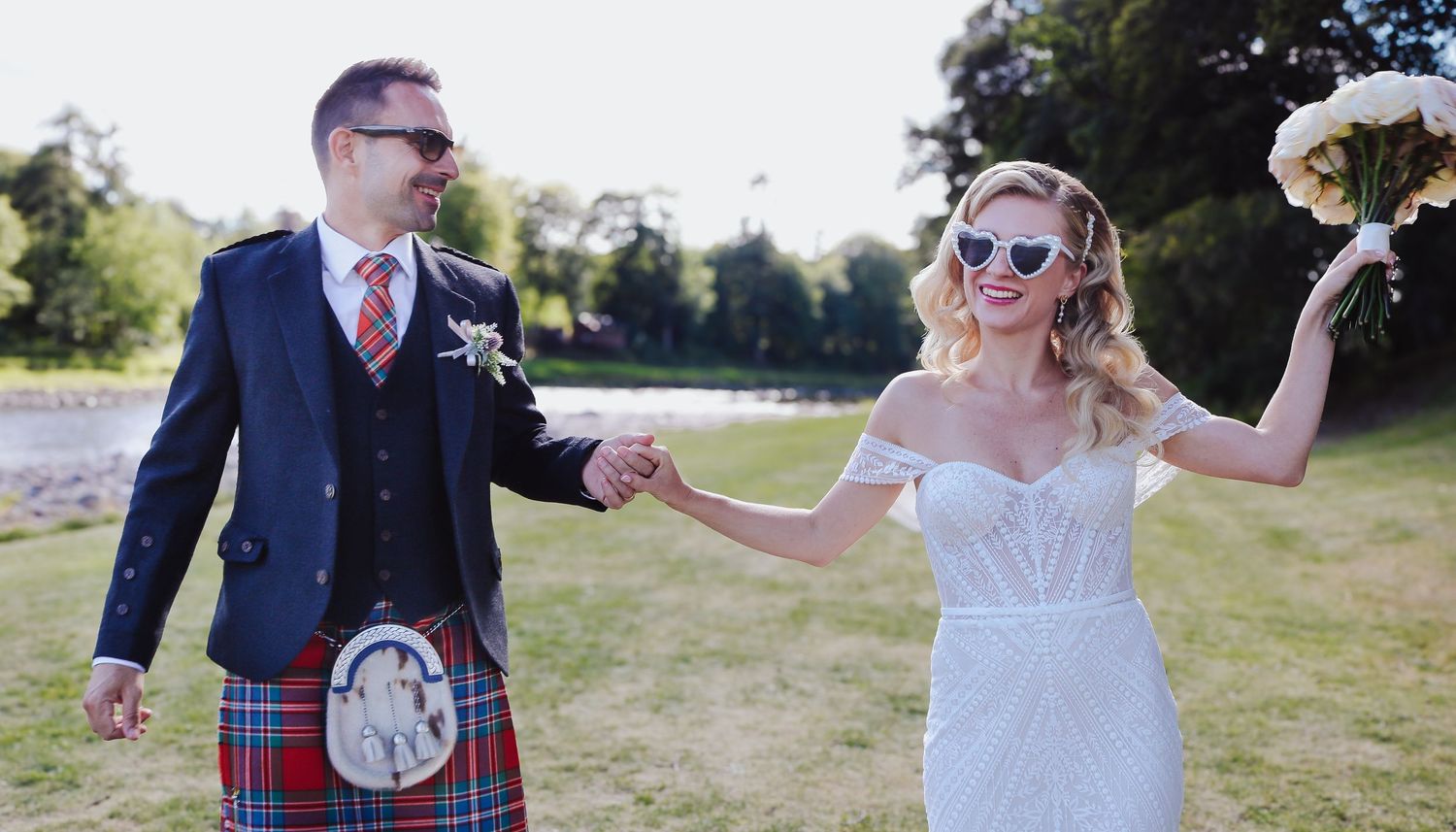 A couple in wedding attire celebrates outdoors, with the groom wearing a traditional Scottish kilt and dark jacket.