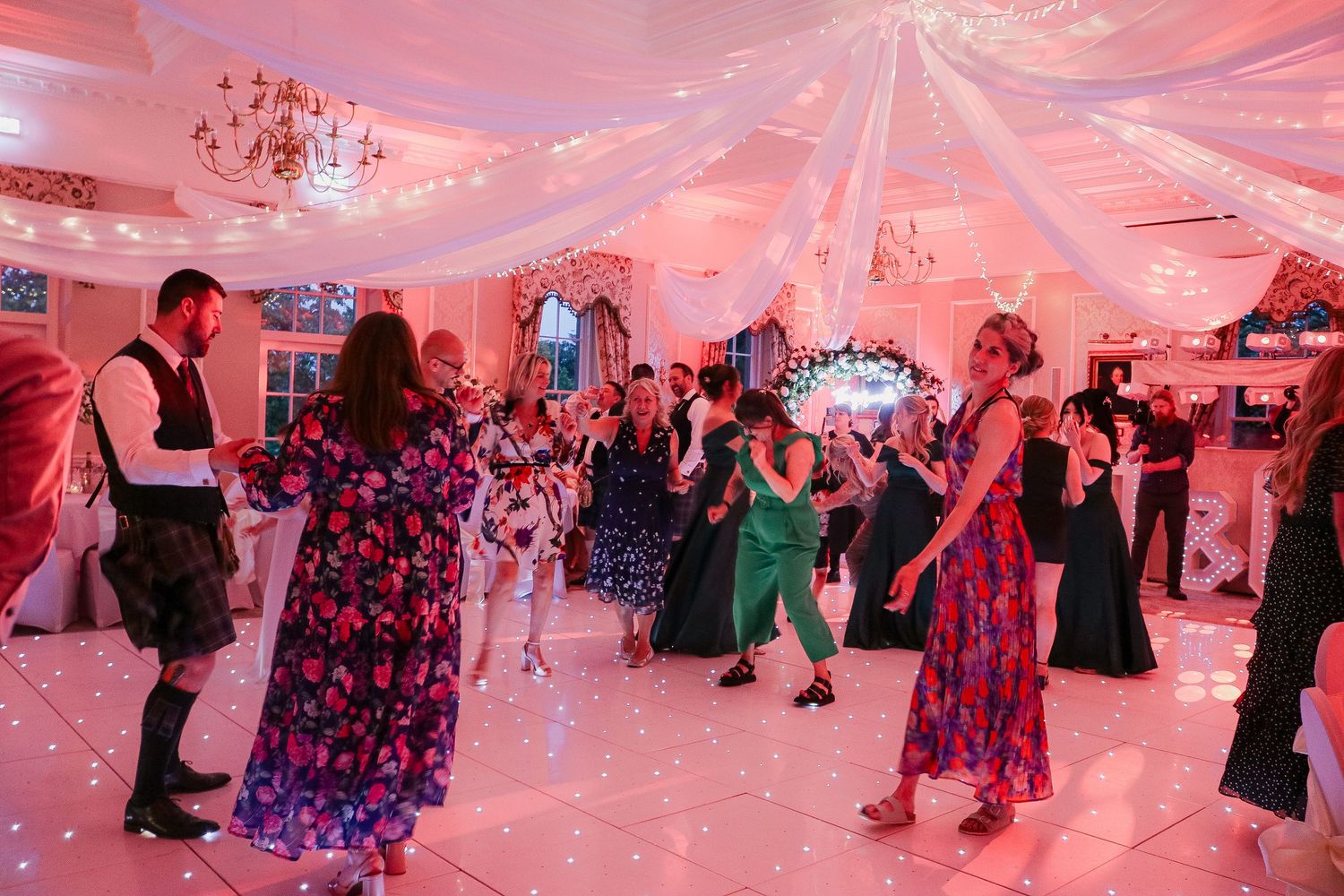 Guests dancing at an elegant wedding reception with pink lighting, draped ceilings, and a white dance floor.