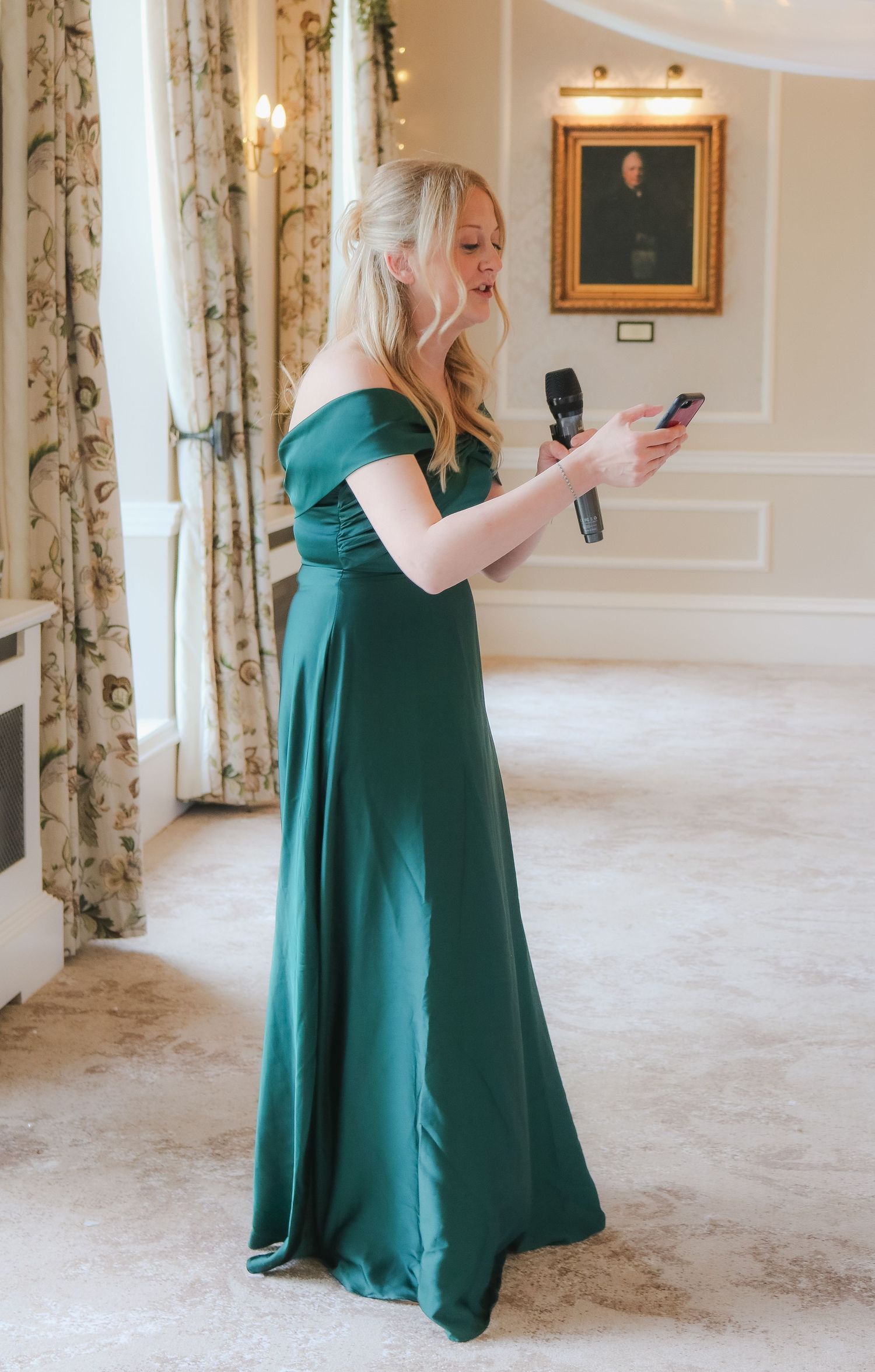 A woman in an elegant emerald green off-shoulder gown holds a microphone and phone while speaking at a formal event.