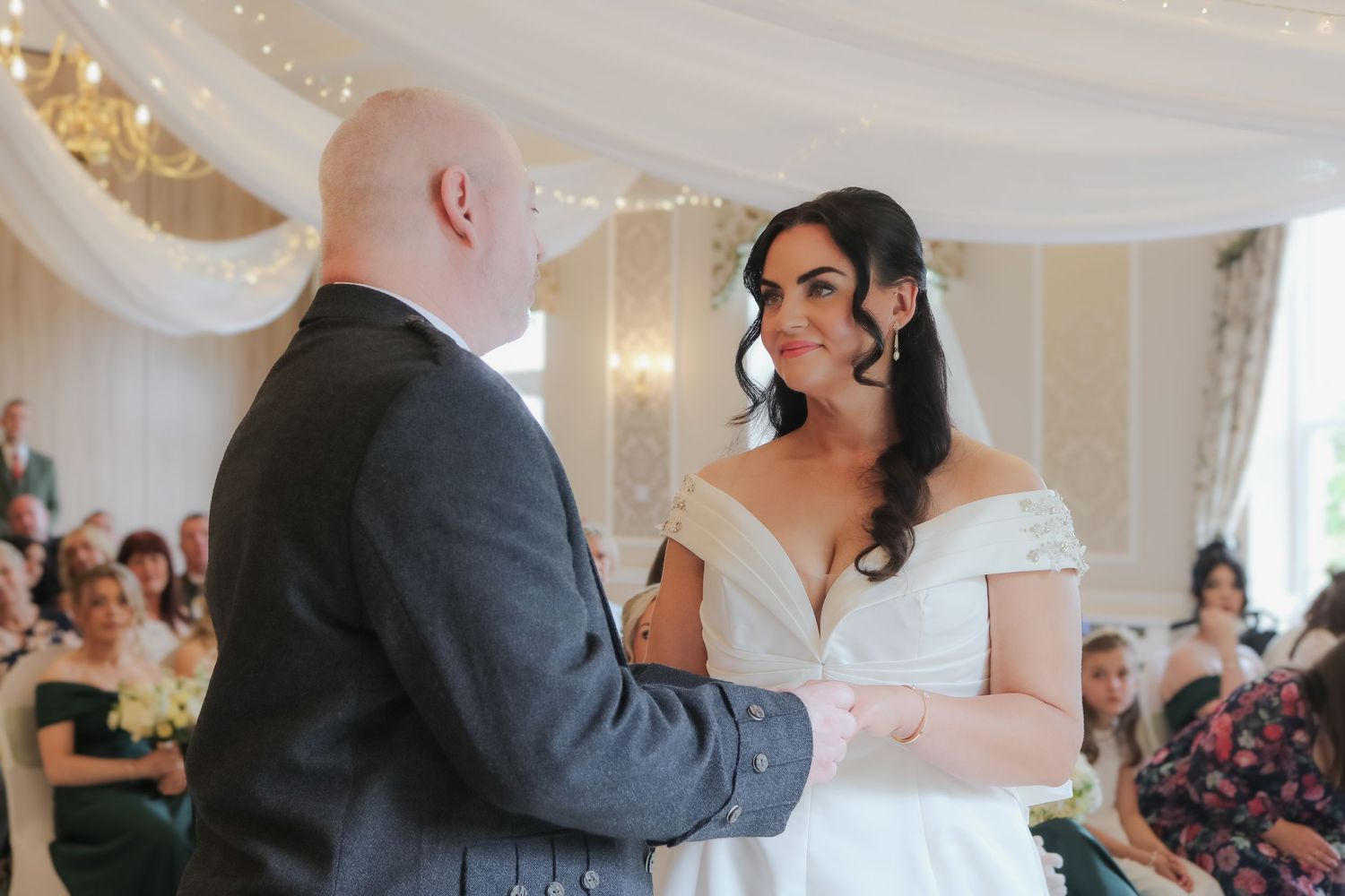 Bride and groom exchanging vows at elegant indoor wedding ceremony with fairy lights and draped white fabric.