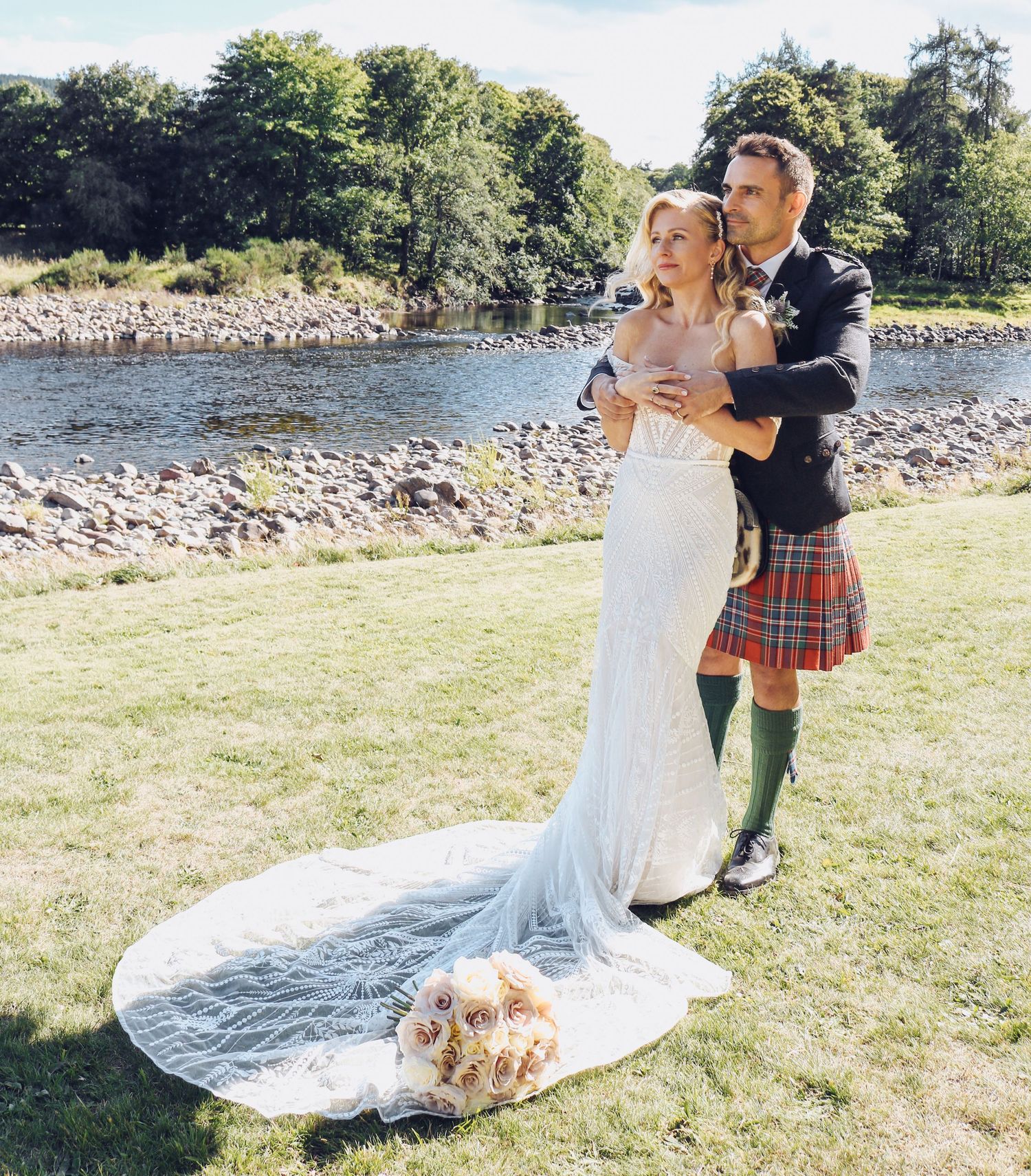 A couple embraces by a scenic riverside on their wedding day, with the groom wearing a traditional Scottish kilt.