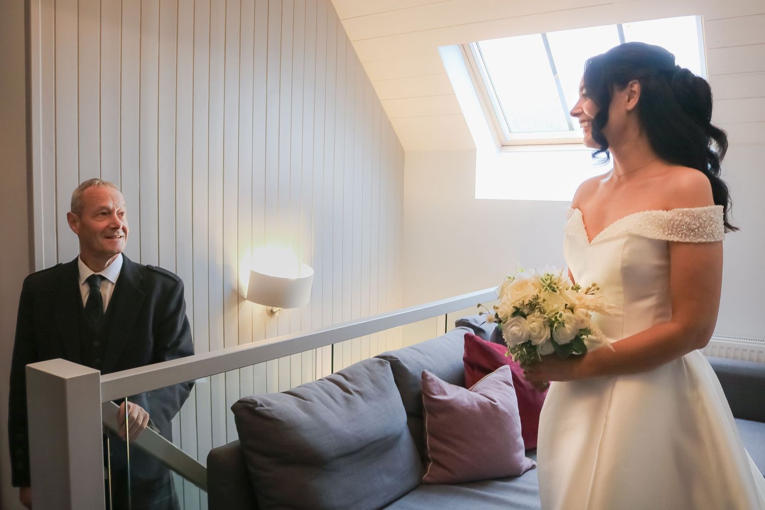 A bride in a white wedding dress stands in a room with white paneled walls and natural light streaming through windows.