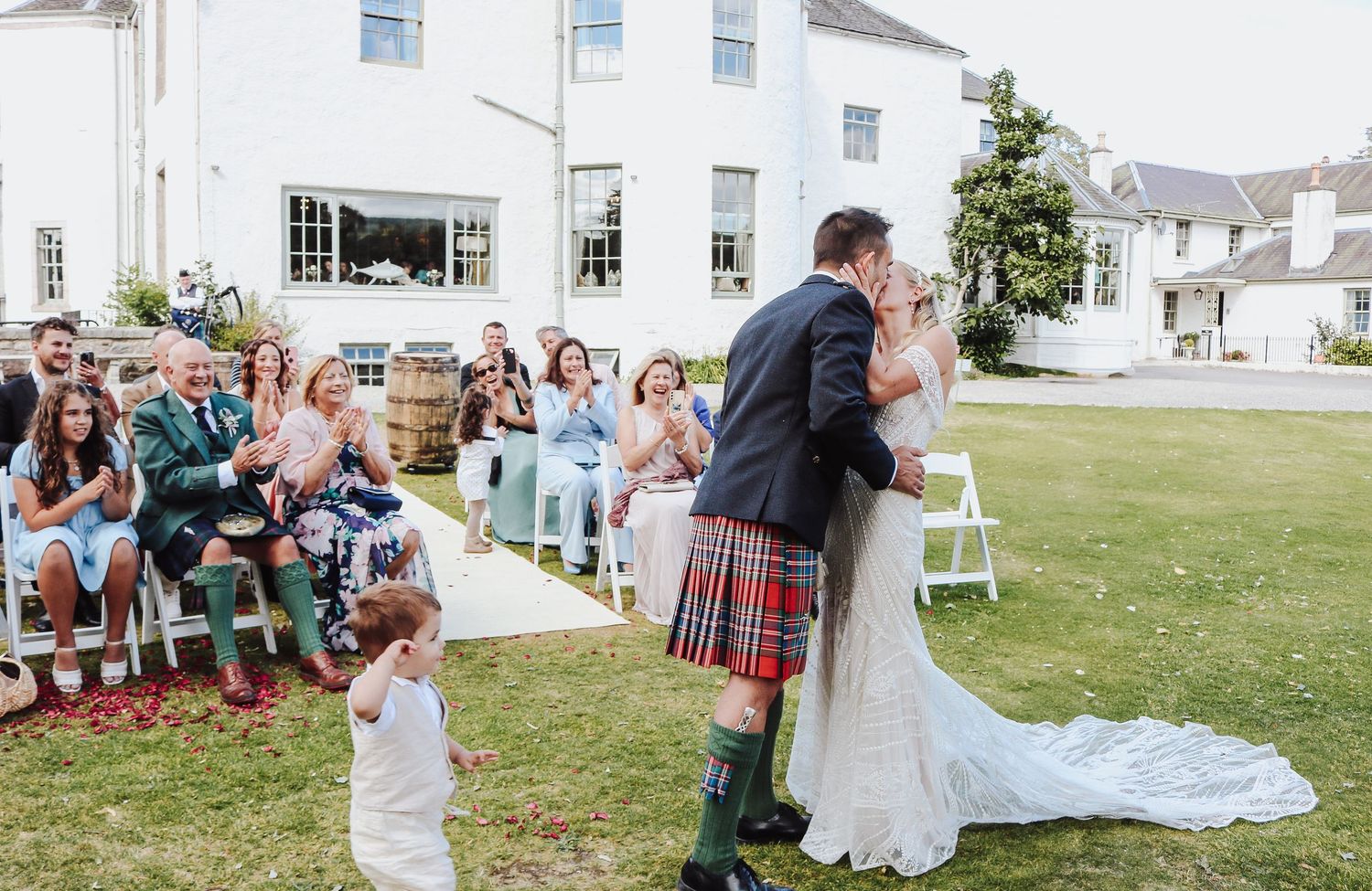 A couple shares a romantic kiss during an outdoor wedding ceremony on a grassy lawn with guests seated nearby.