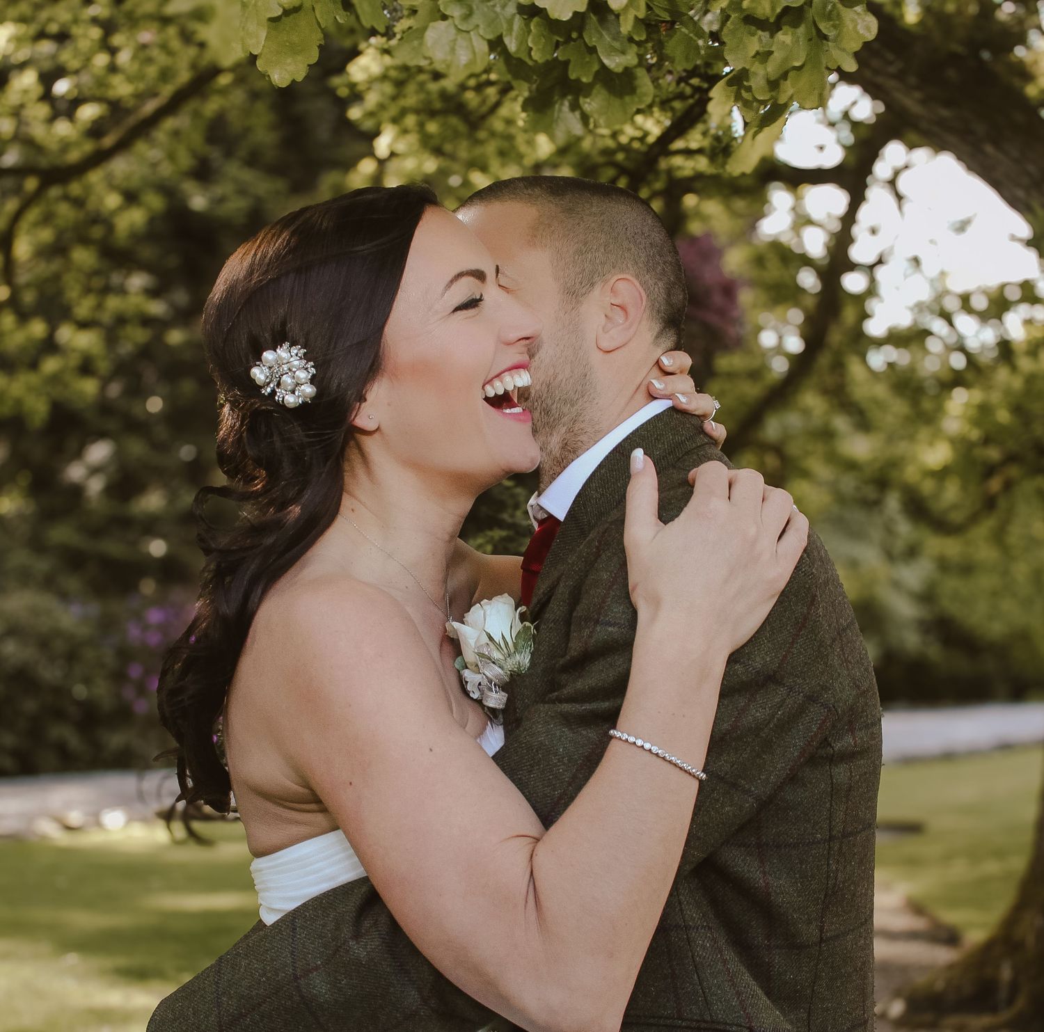 A joyful wedding couple sharing a candid laughing moment together outdoors with green foliage in the background.