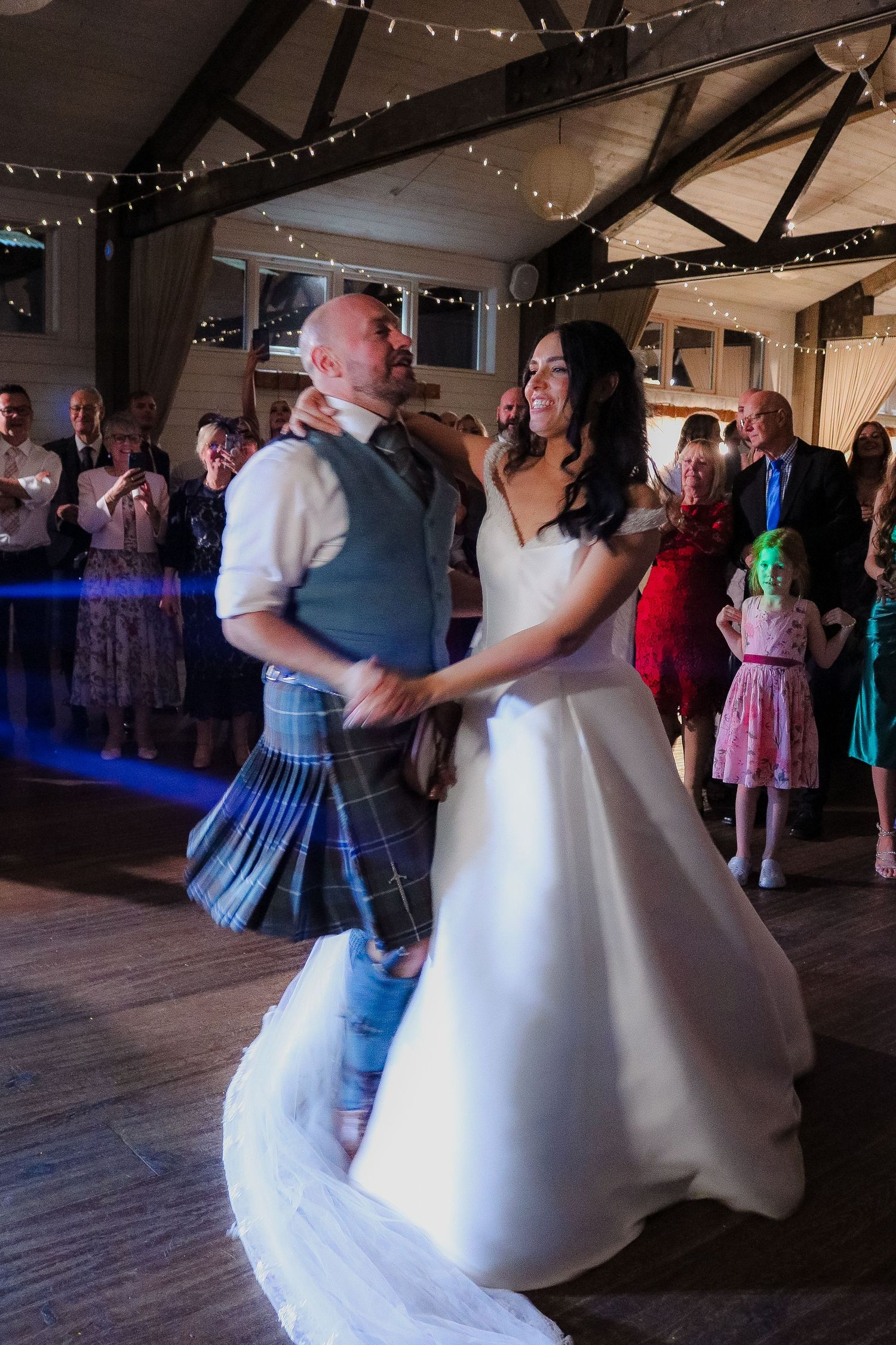 A couple dances at a wedding reception in a rustic venue decorated with string lights, with the groom wearing a traditional kilt.