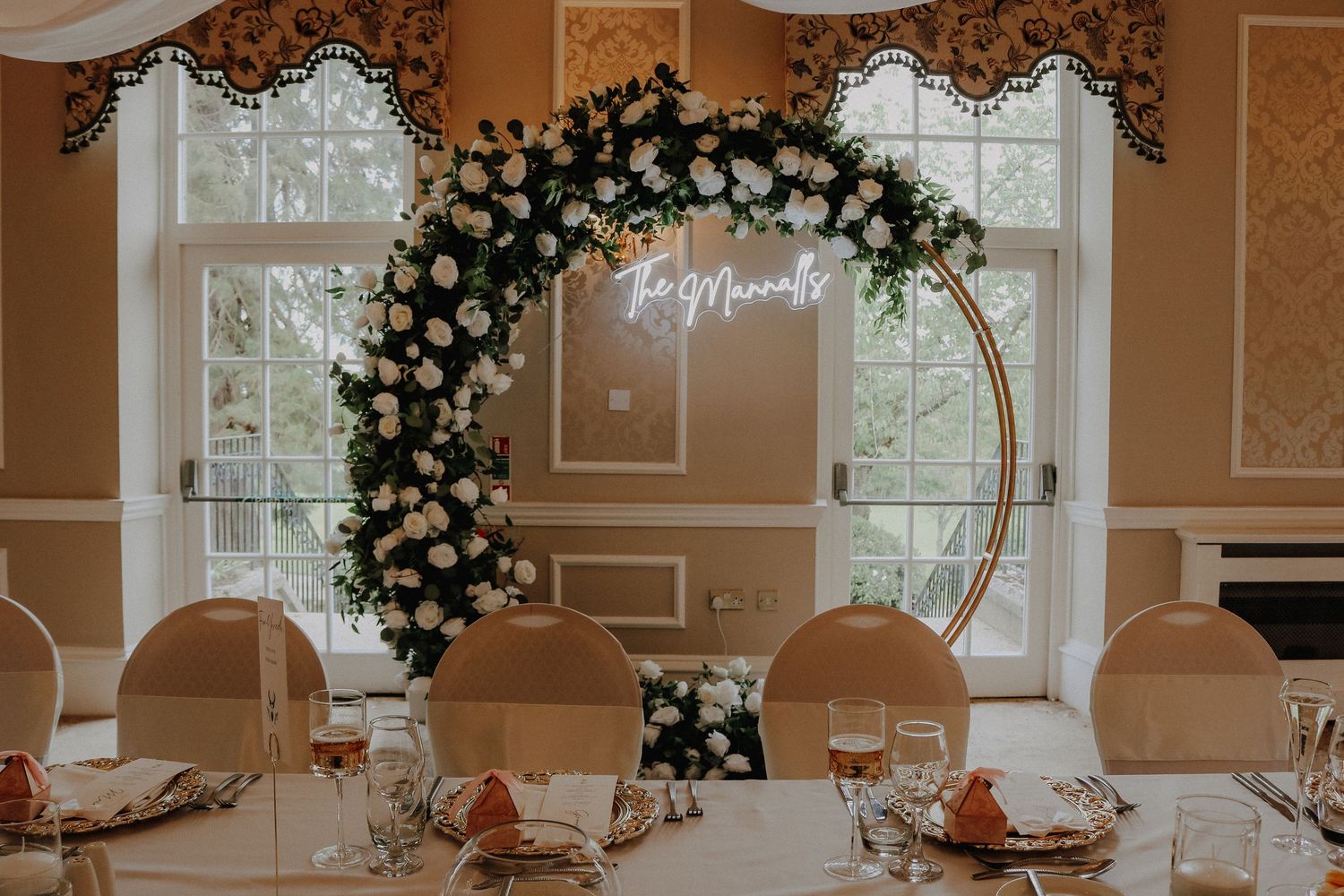 Elegant wedding reception with a floral hoop arch featuring white roses, greenery, and a neon sign above a decorated banquet table.