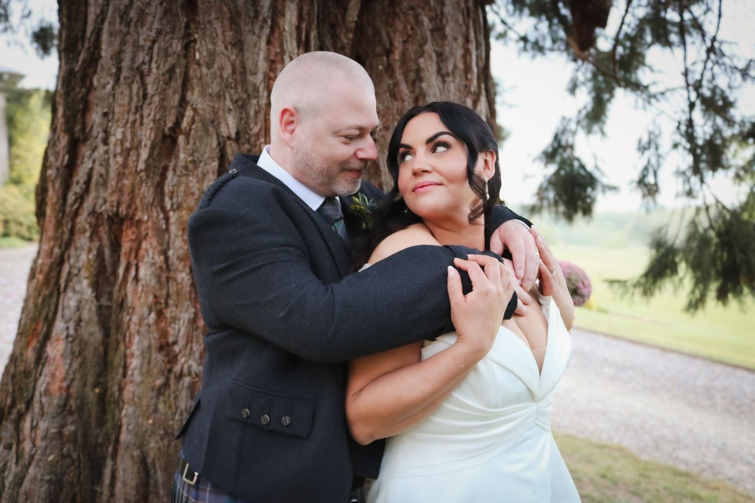 A bride and groom embrace romantically against a large tree trunk outdoors on their wedding day.