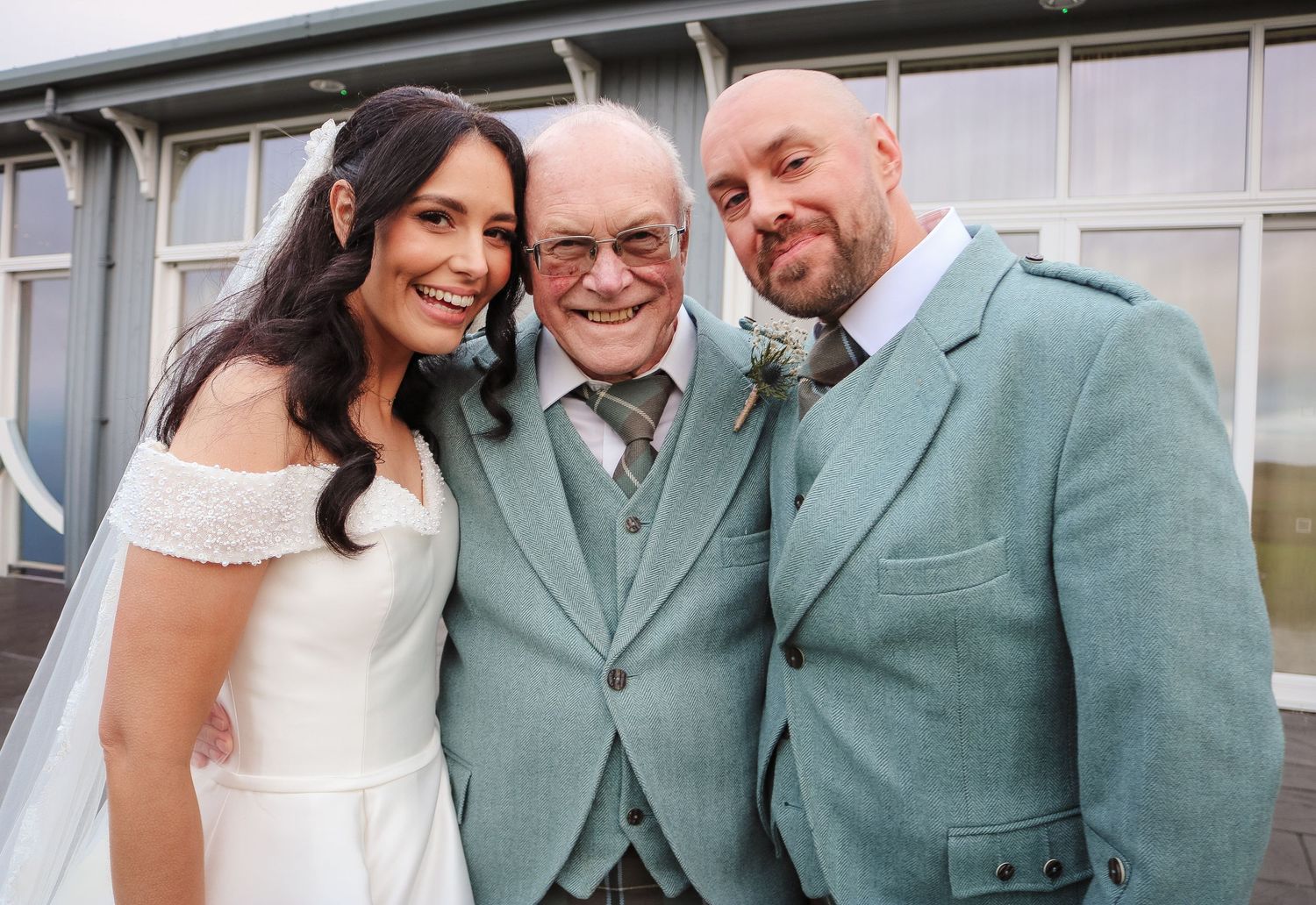 A group of people in formal attire pose together at a wedding celebration wearing sage green and white outfits.